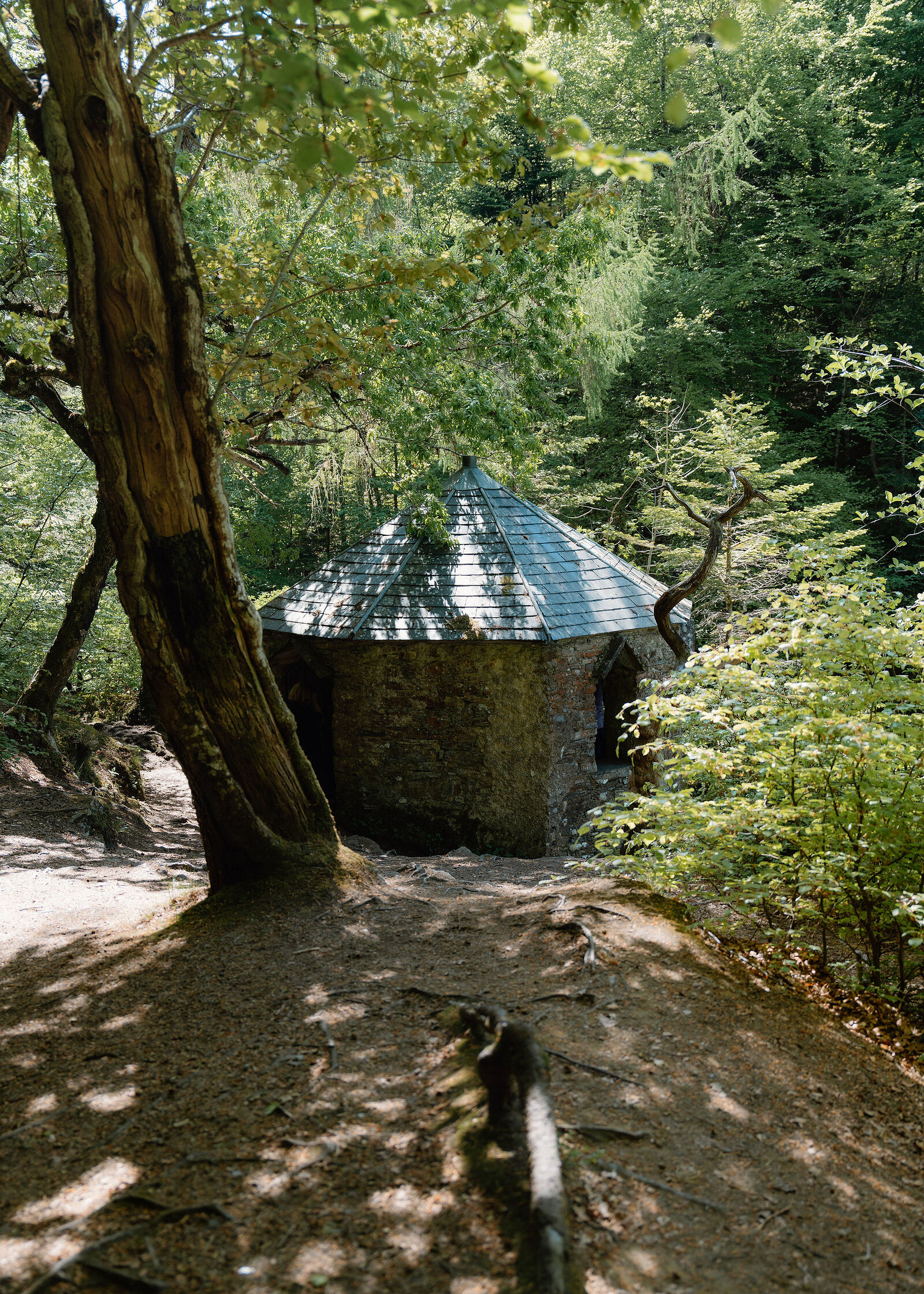 A small stone summerhouse with a conical slate roof among trees, a dappled earth path leading to it.