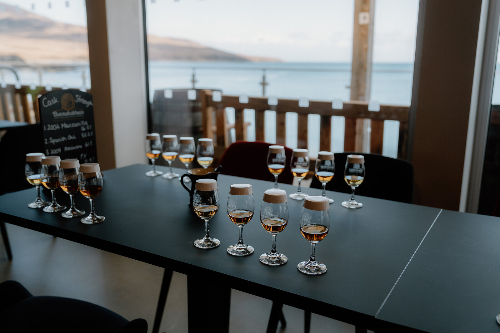 Tasting glasses of whisky line a table before a window overlooking the sea.