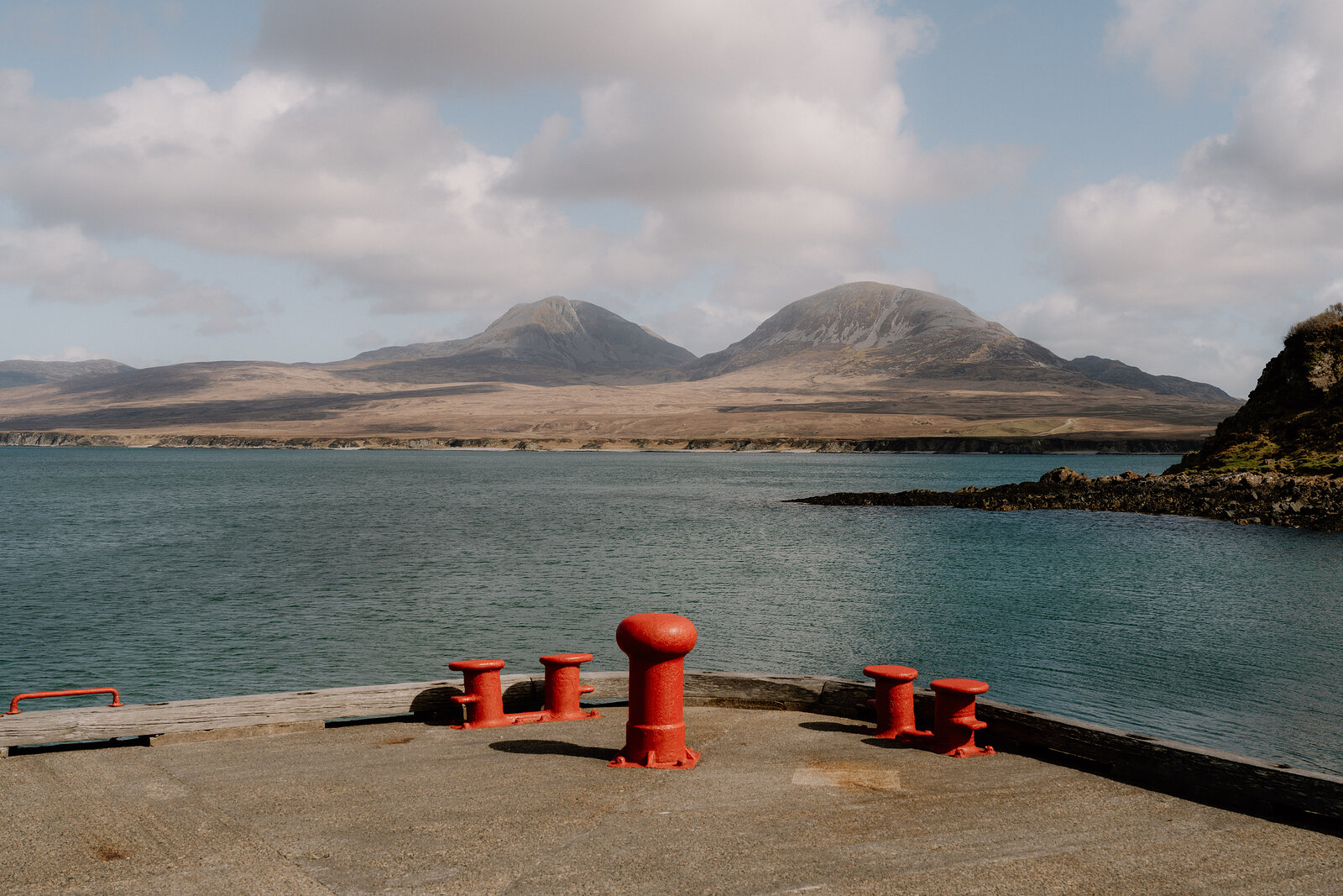 Red mooring bollards line a stone pier before distant mountains.