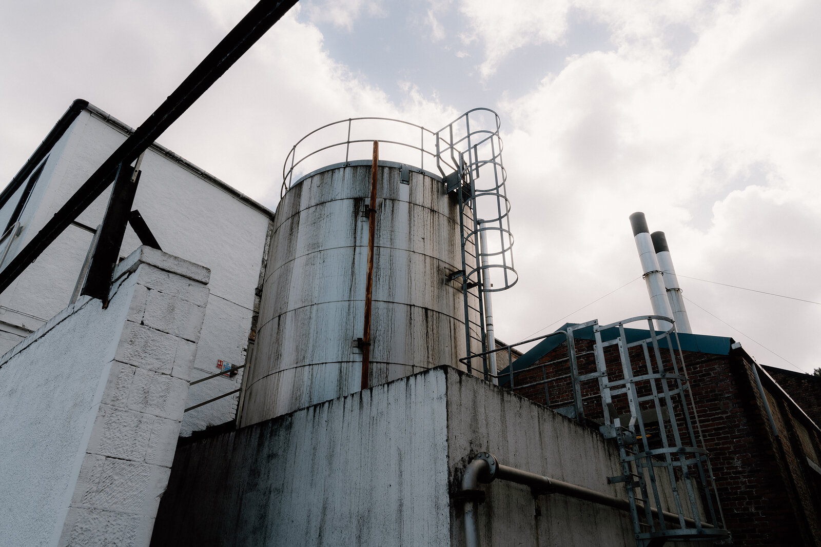 A metal storage tank with a ladder rises beside distillery buildings.