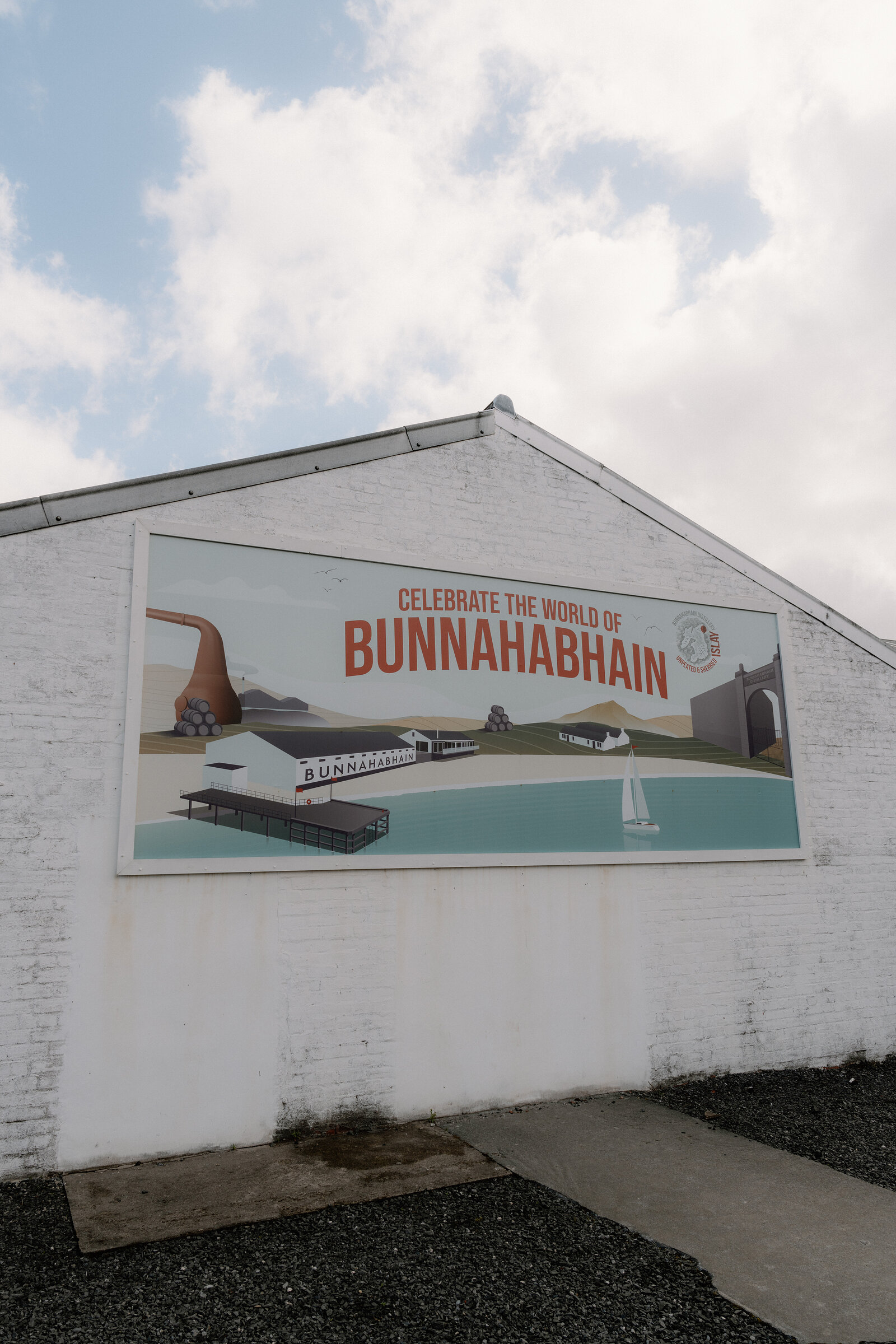 An illustrated mural reads "Celebrate the World of Bunnahabhain" on a distillery gable wall.
