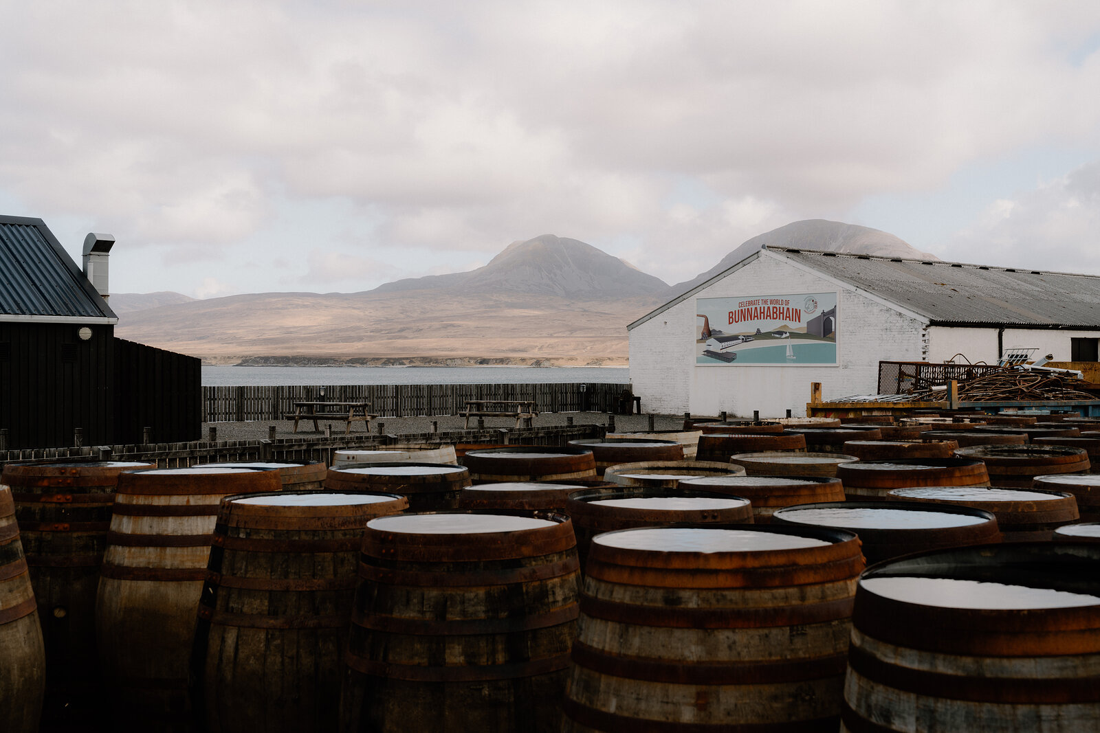Whisky casks stand before Bunnahabhain's mural-painted gable wall beneath distant hills.