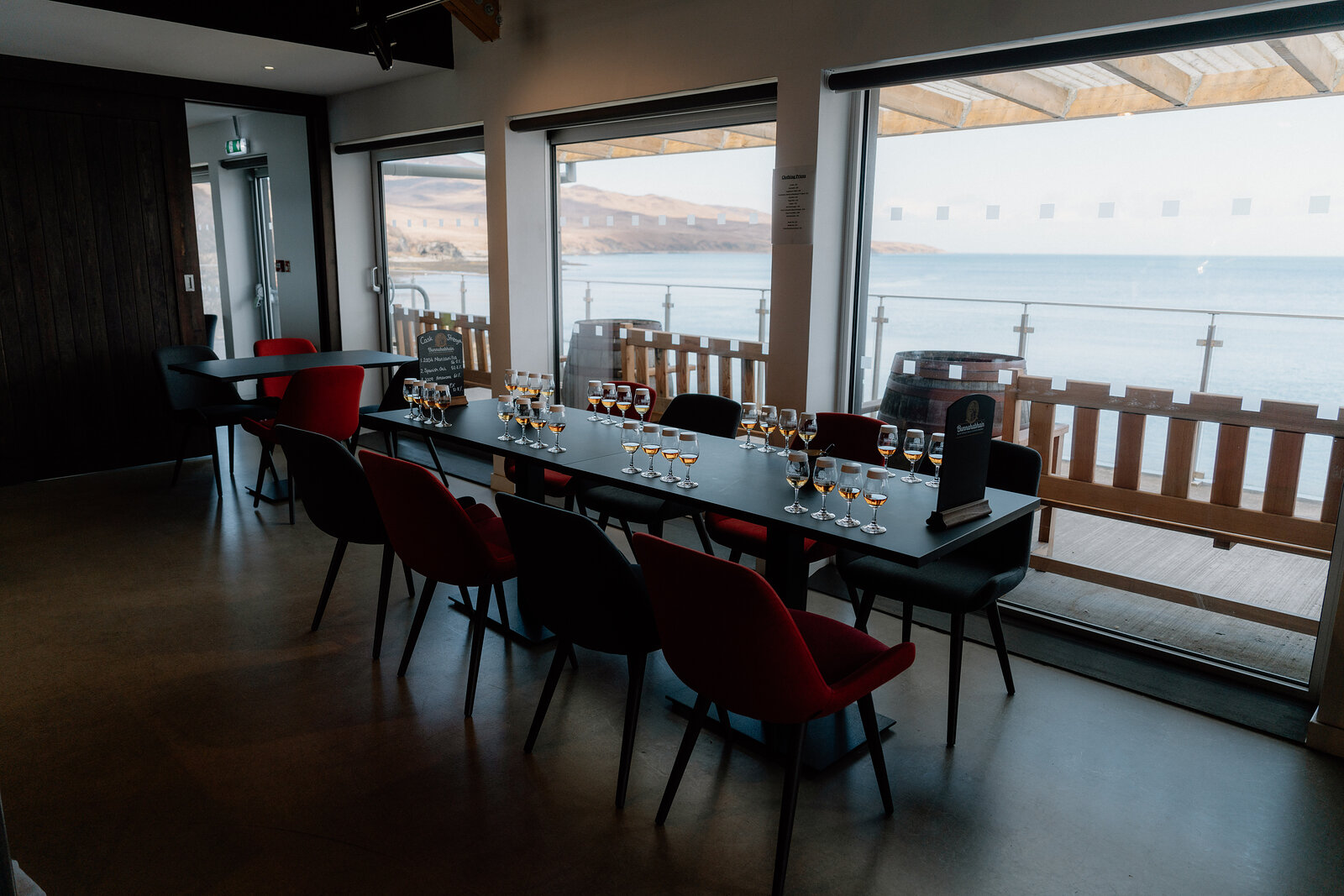 Red chairs line a long tasting table set with whisky glasses before a window overlooking the sea.