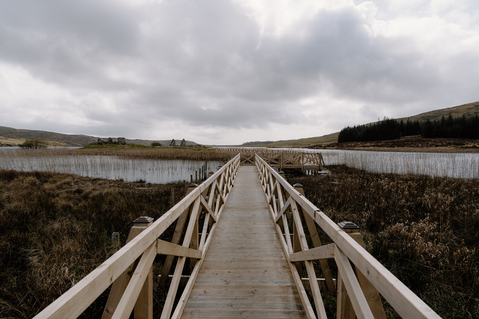 A wooden boardwalk bridge crosses a reed-lined loch toward distant buildings.