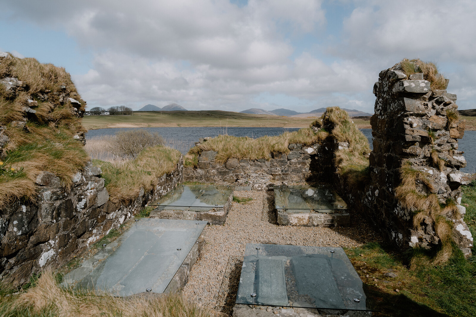 Glass-covered grave slabs lie within the ruined chapel walls at Finlaggan, a loch beyond.