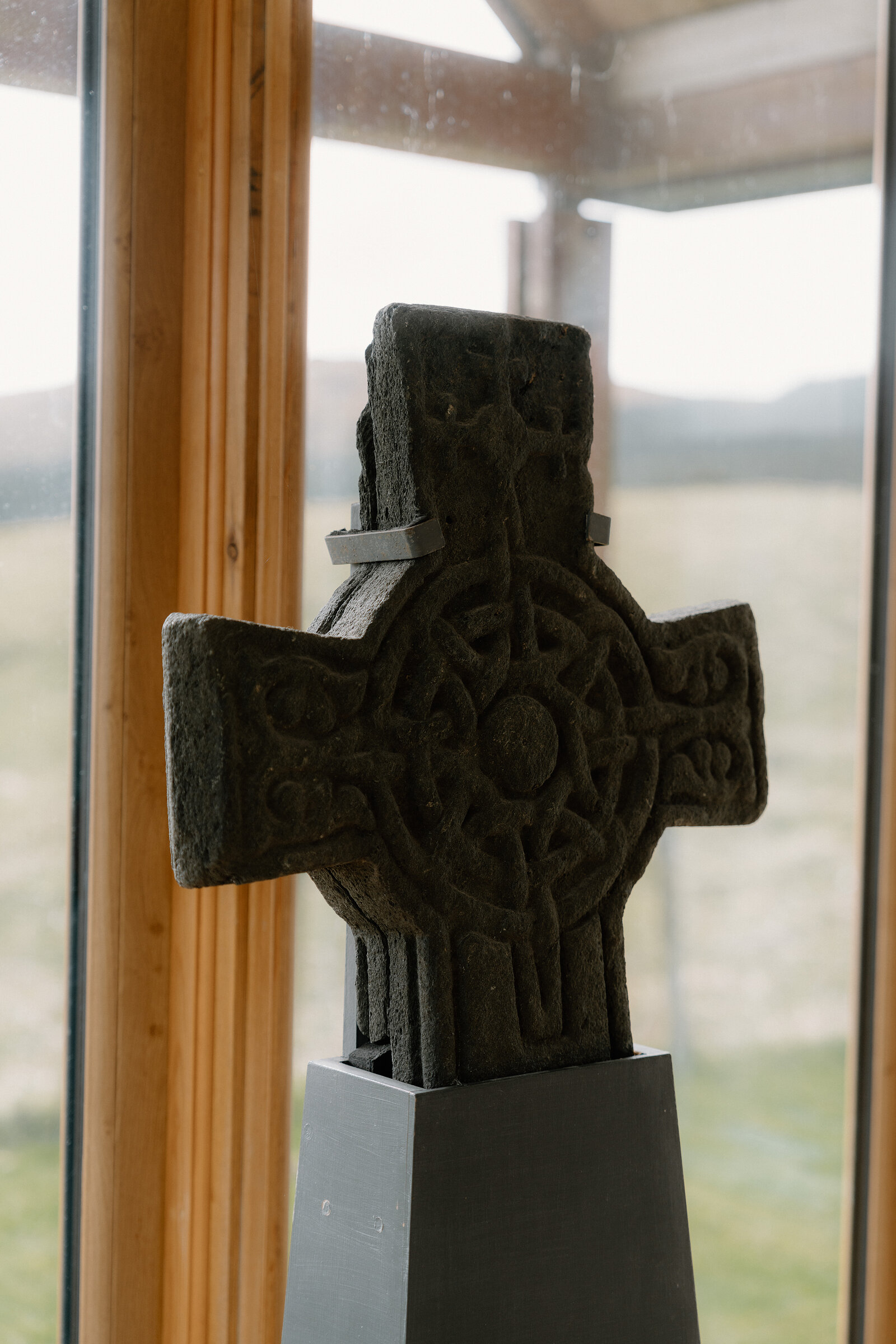A carved stone Celtic cross stands on a plinth beside a window overlooking open land.