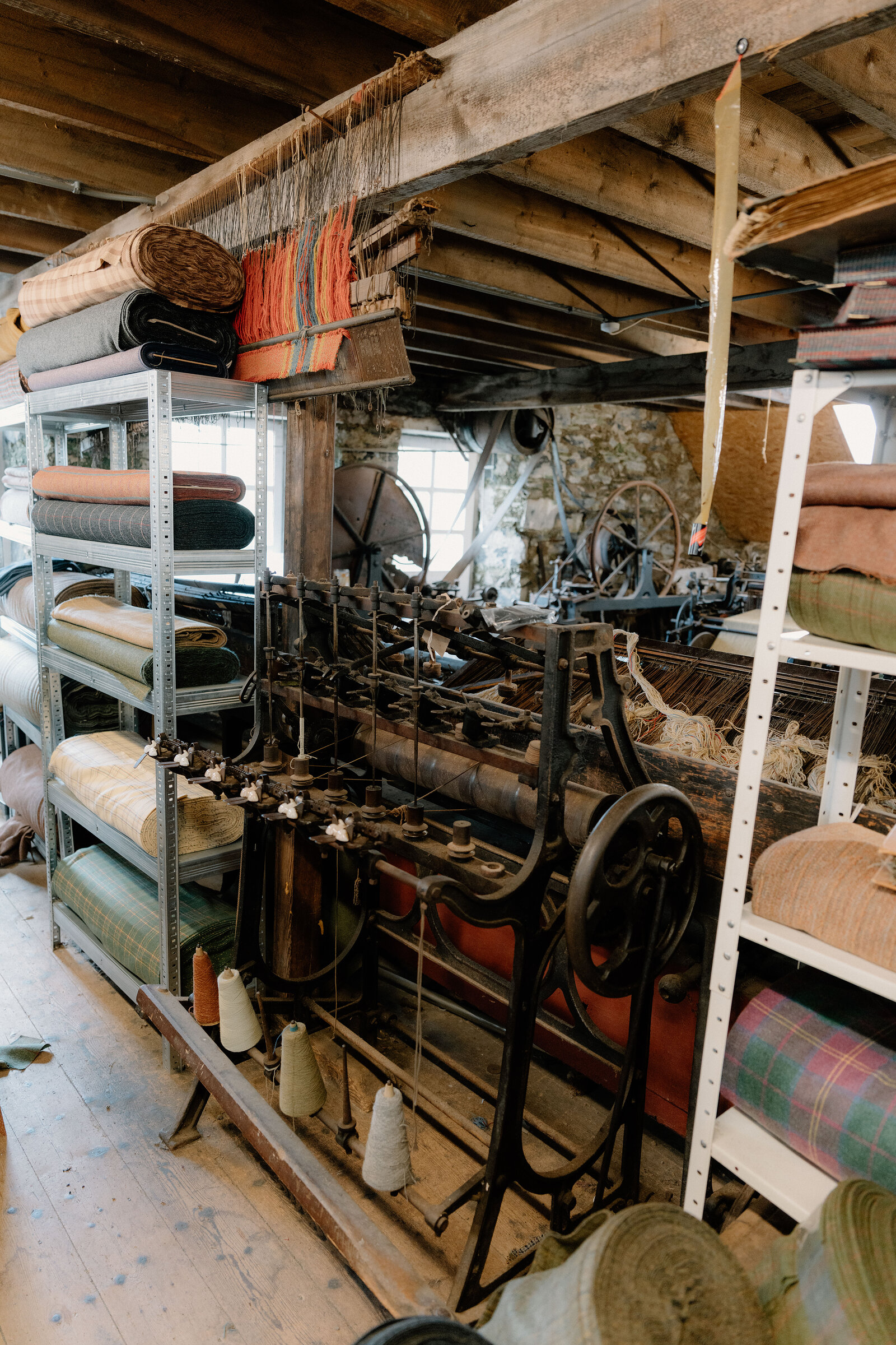 Rolls of tartan fabric and a working loom fill a stone-walled weaving workshop.