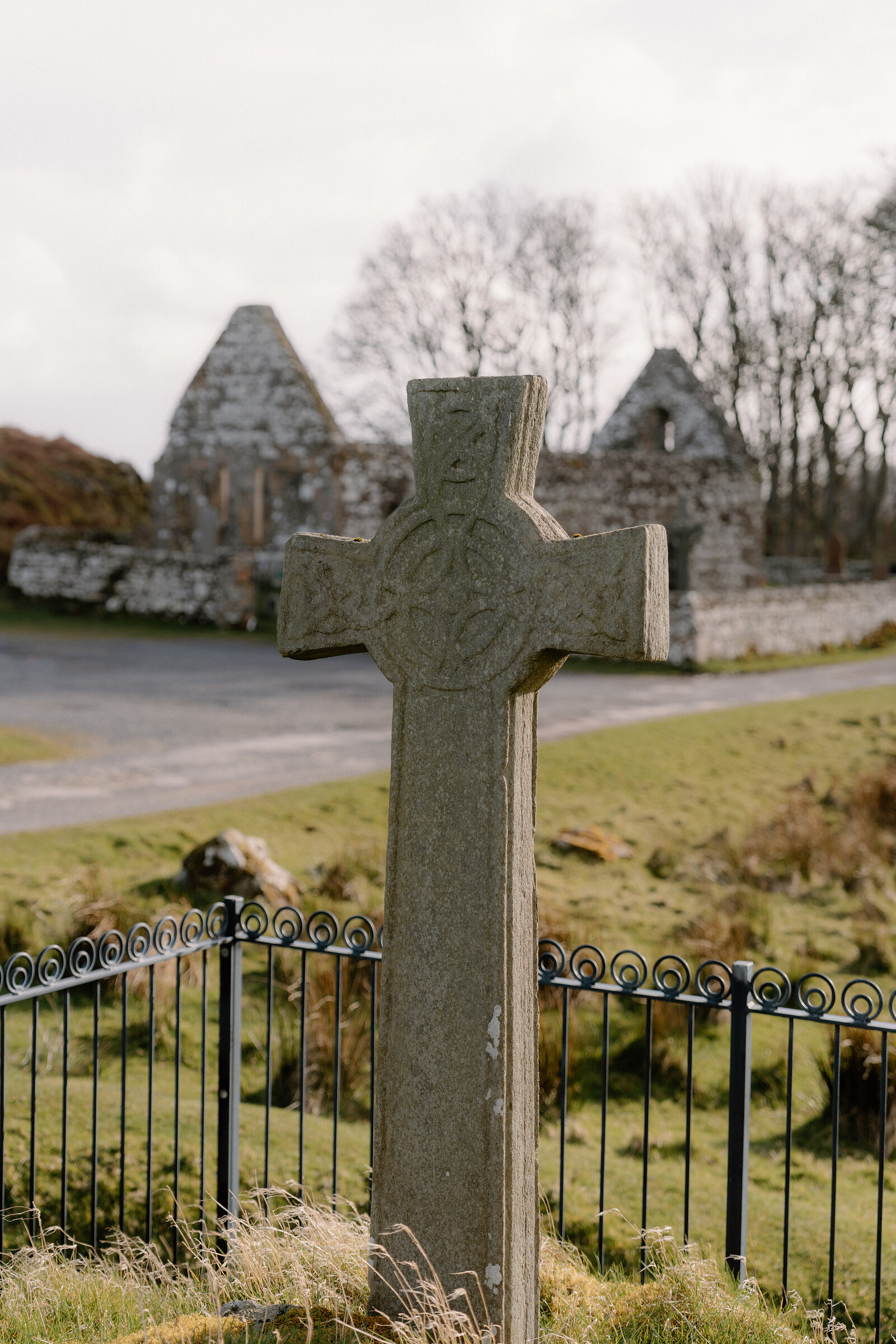 A carved stone cross stands before the ruined gable of a stone chapel.
