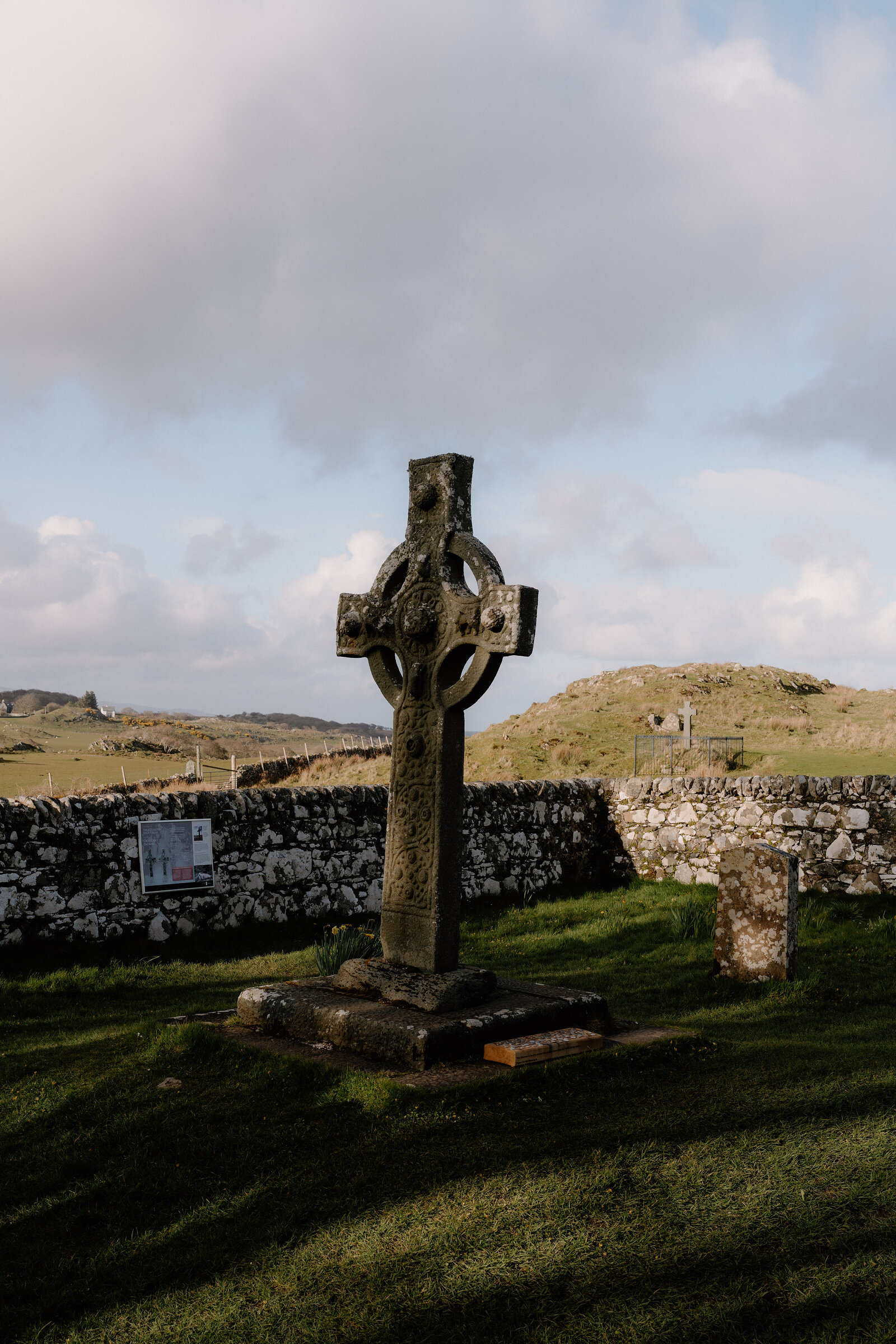 A weathered carved cross stands in a churchyard beside a low stone wall and distant hill.