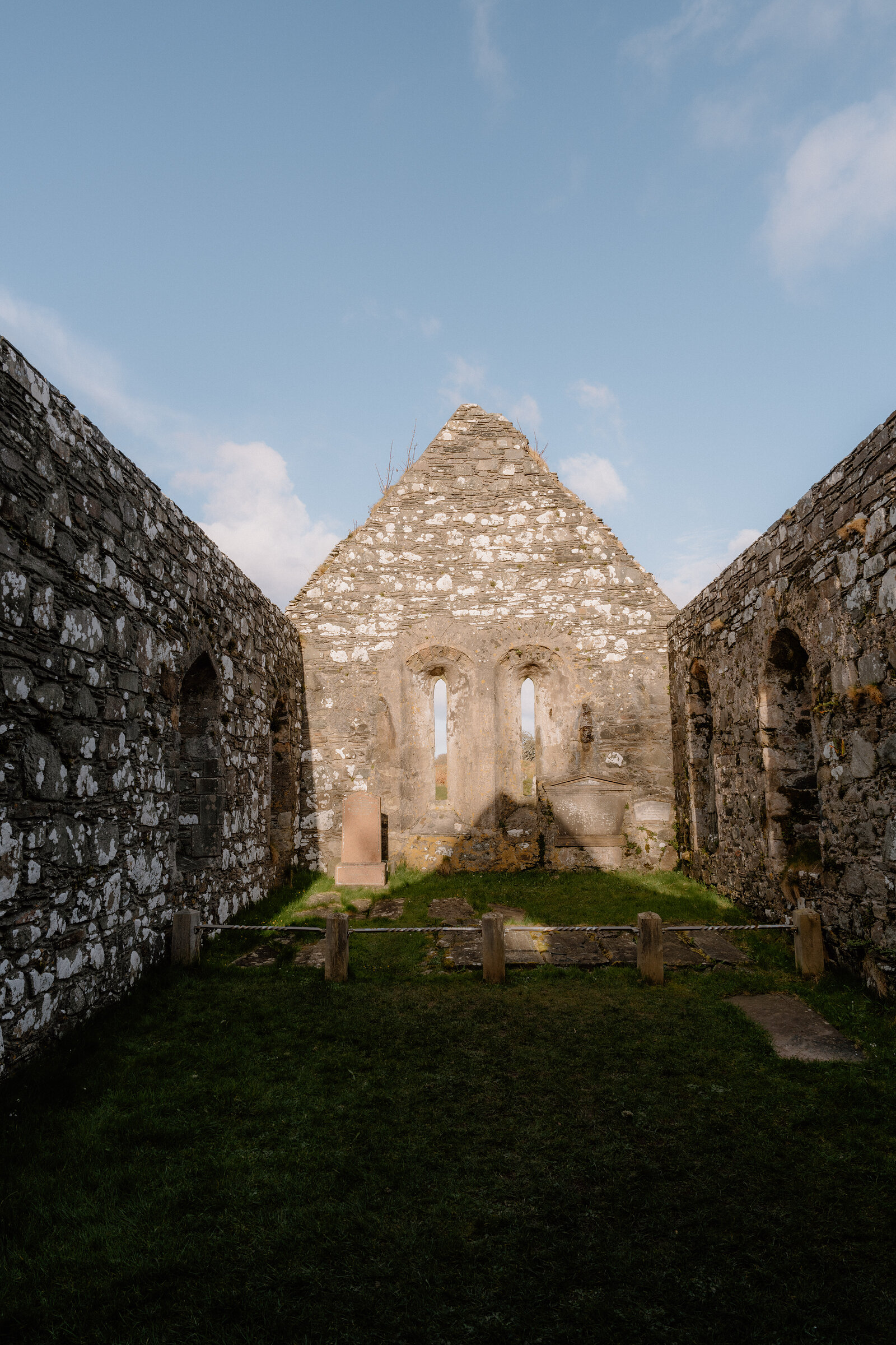 Pointed gothic windows pierce the ruined gable wall of a roofless stone chapel.