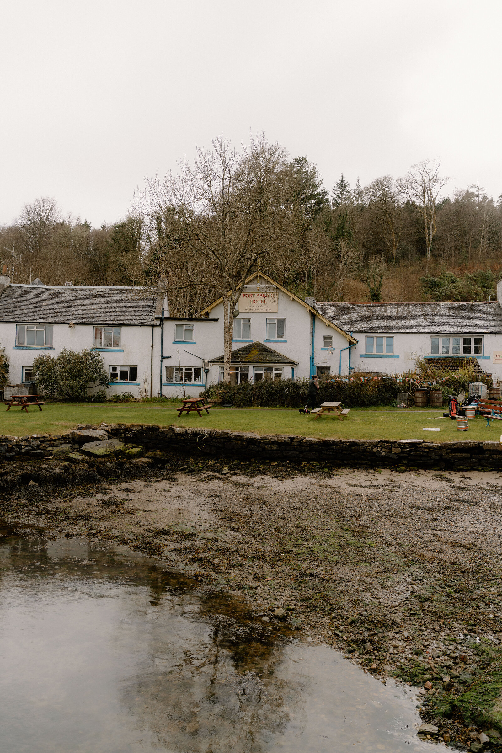 A whitewashed hotel building faces a grass lawn with picnic tables that runs down to the shore of a sea loch, wooded hills behind.