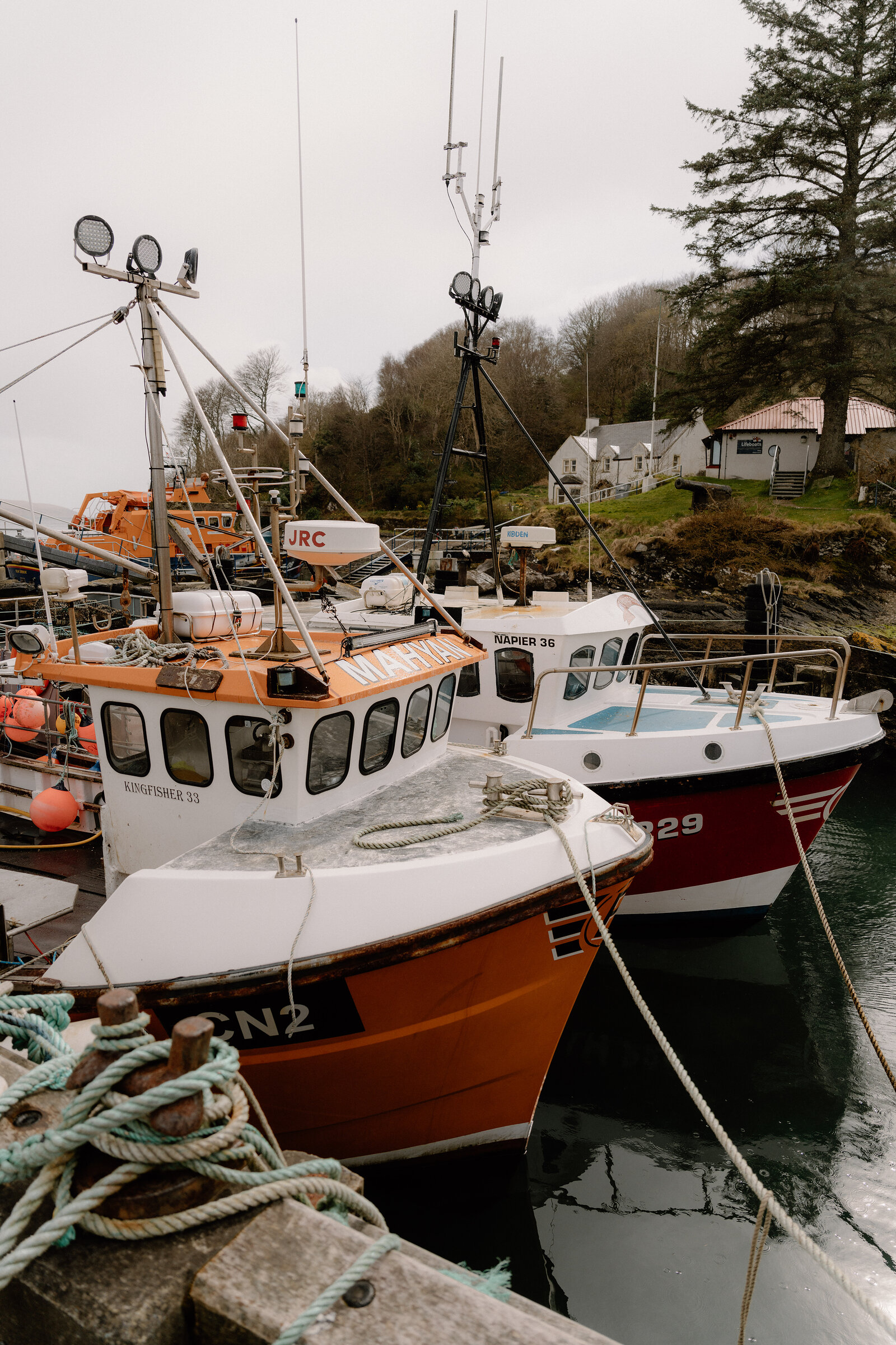 Orange and white fishing boats named Kingfisher 33 and Kingfisher 36 are moored together in a harbour beside a house on a wooded hill.
