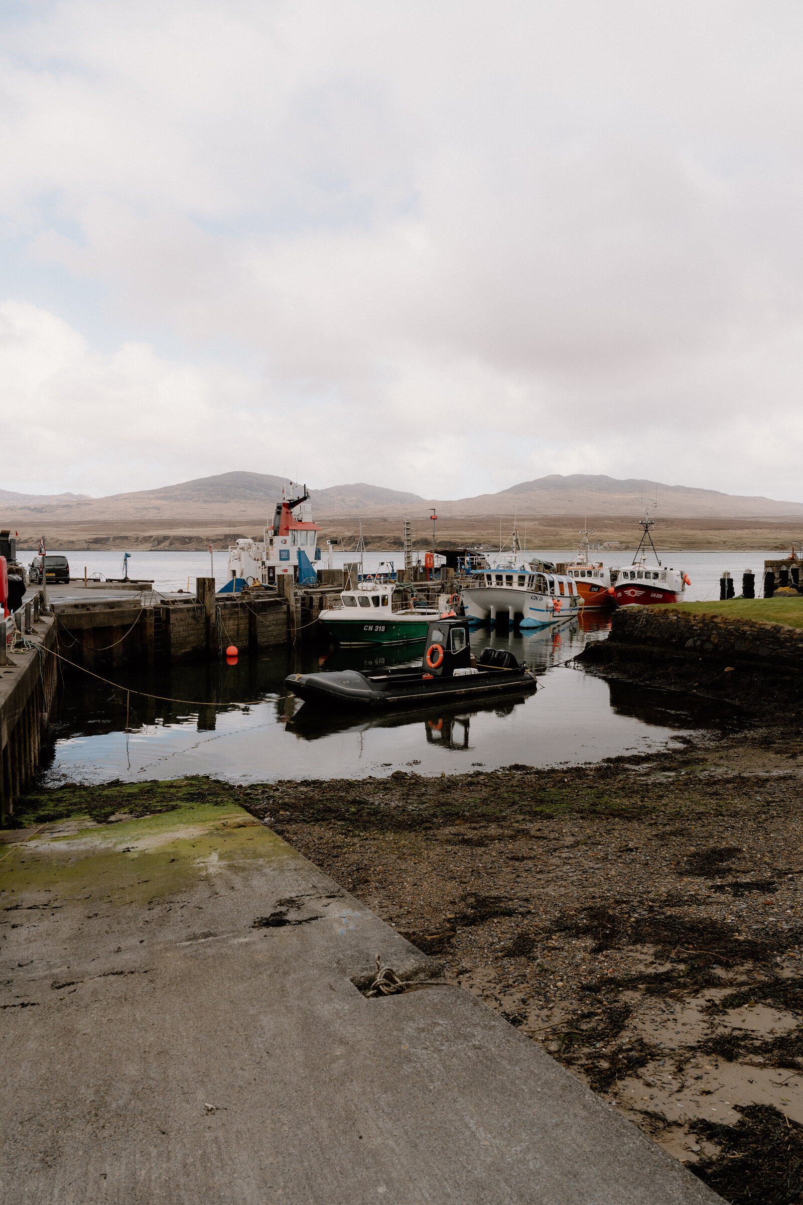 Fishing boats and a small ferry are moored at a stone harbour at low tide, hills rising across the water beyond.