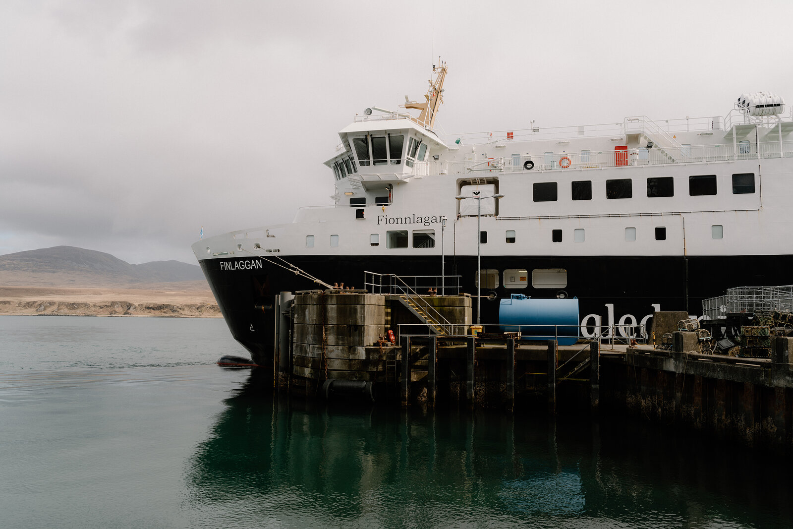 The ferry Fionnlagan is docked beside sandy hills, its hull reflected in the calm water.