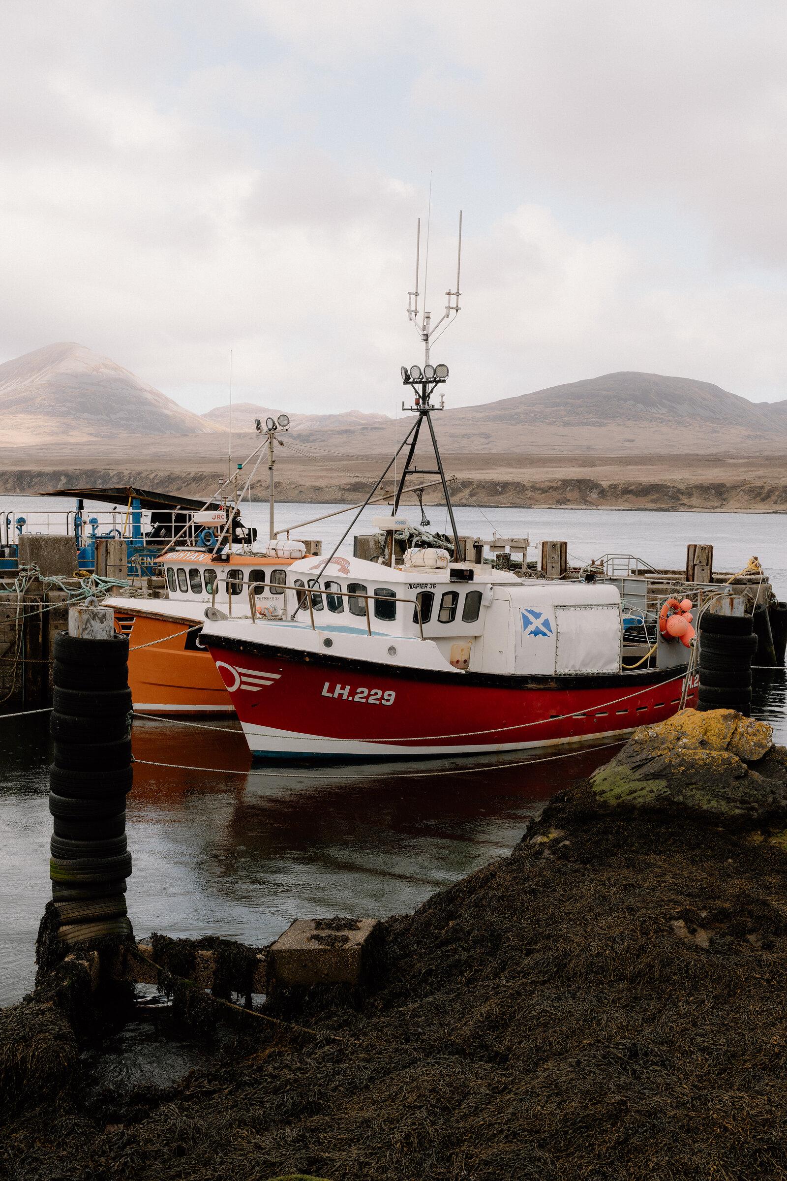 A red and white fishing boat marked LH.229 is moored at a stone harbour beneath distant hills.