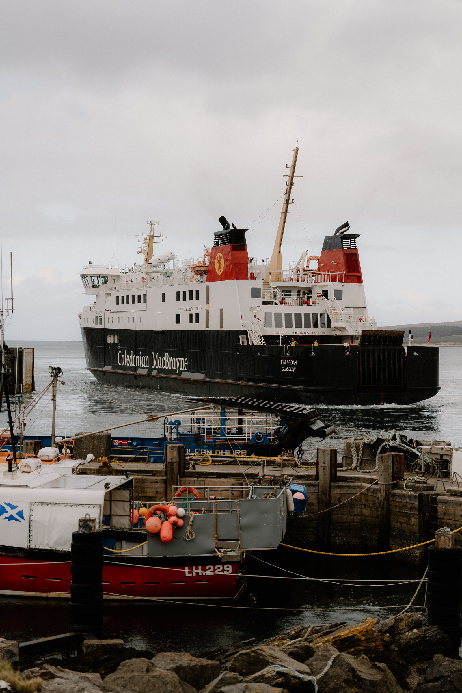 A Caledonian MacBrayne ferry manoeuvres into Port Askaig harbour past fishing boats flying a Saltire flag.