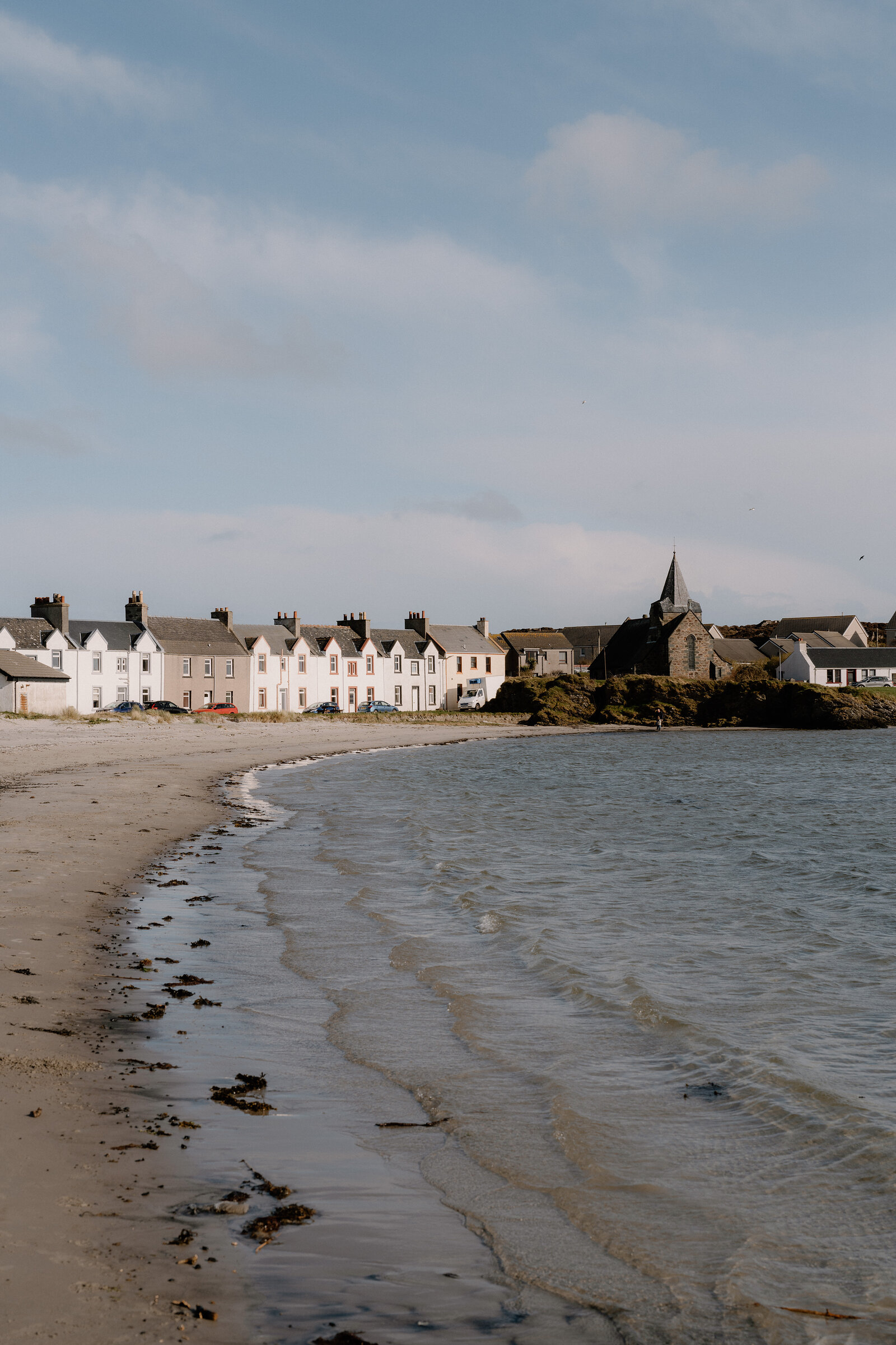 A church spire rises above white terraced houses lining a curved beach in an Islay village.