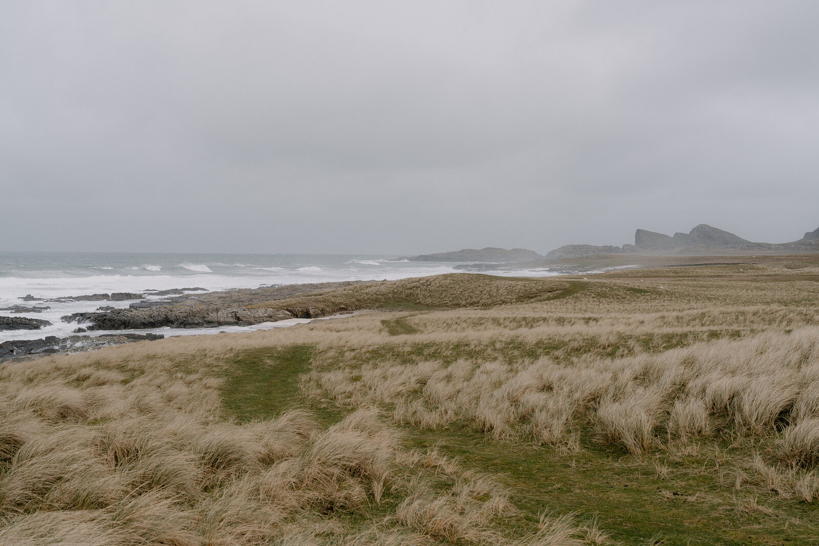 Wind-blown grass dunes lead to a rocky shoreline where waves break beneath a headland.