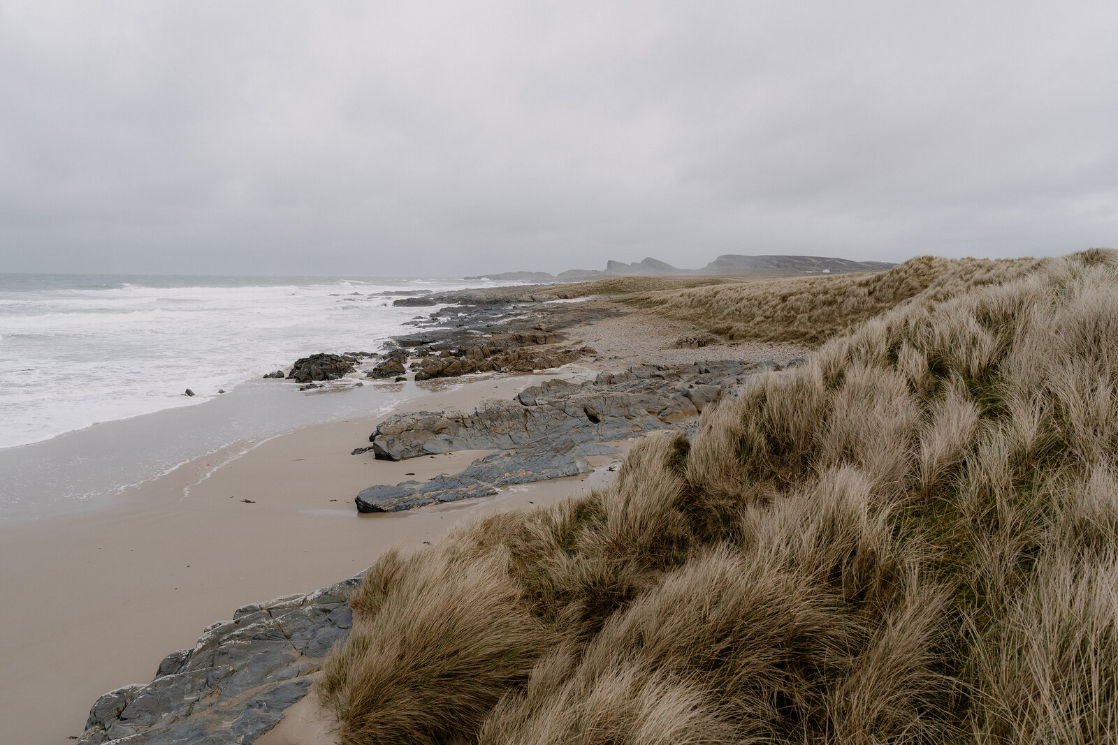 Wind-blown dune grass overlooks a rocky shoreline where waves break beneath a misty headland.