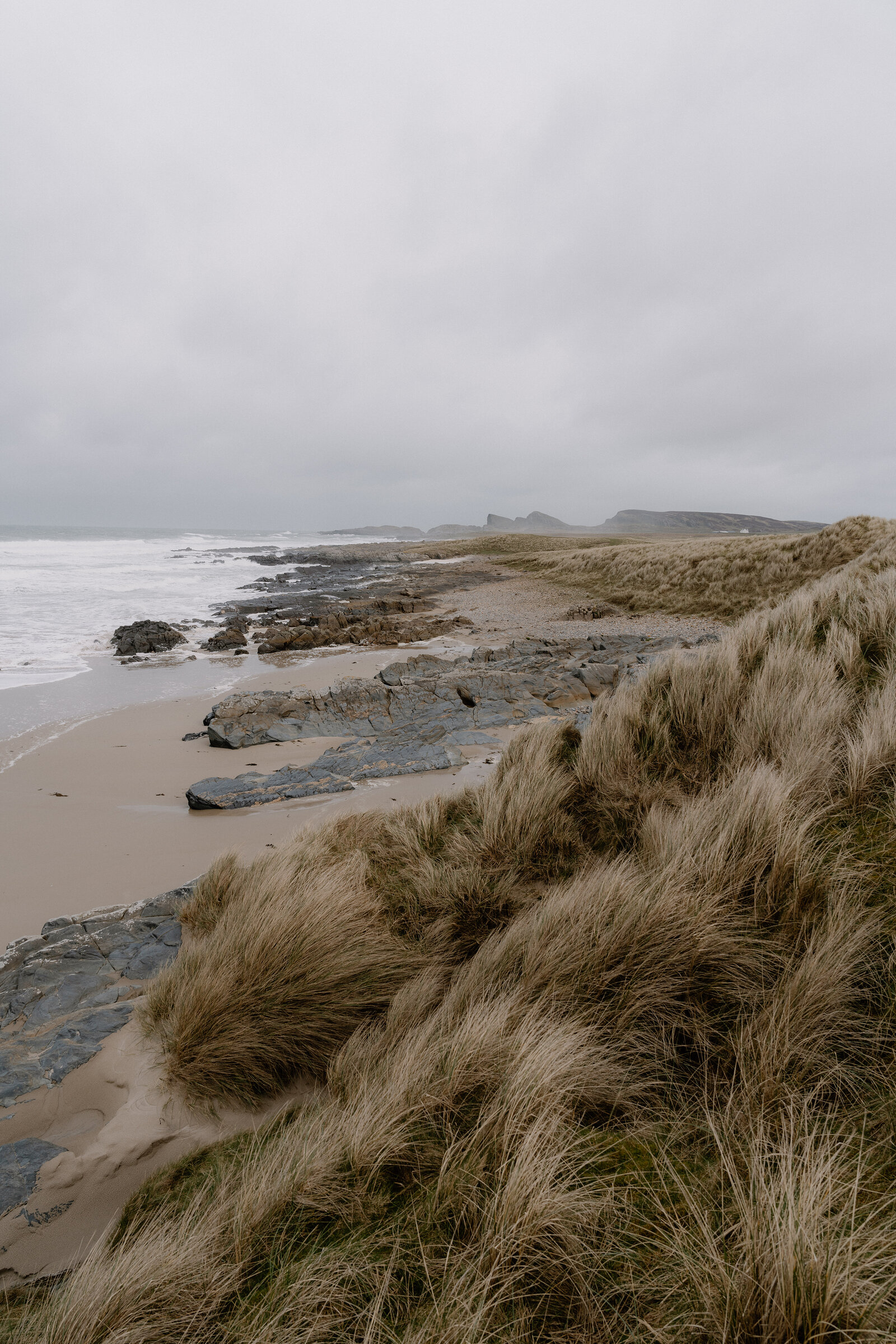 Layered rocks line a beach beneath windswept dunes as waves roll in under a grey sky.