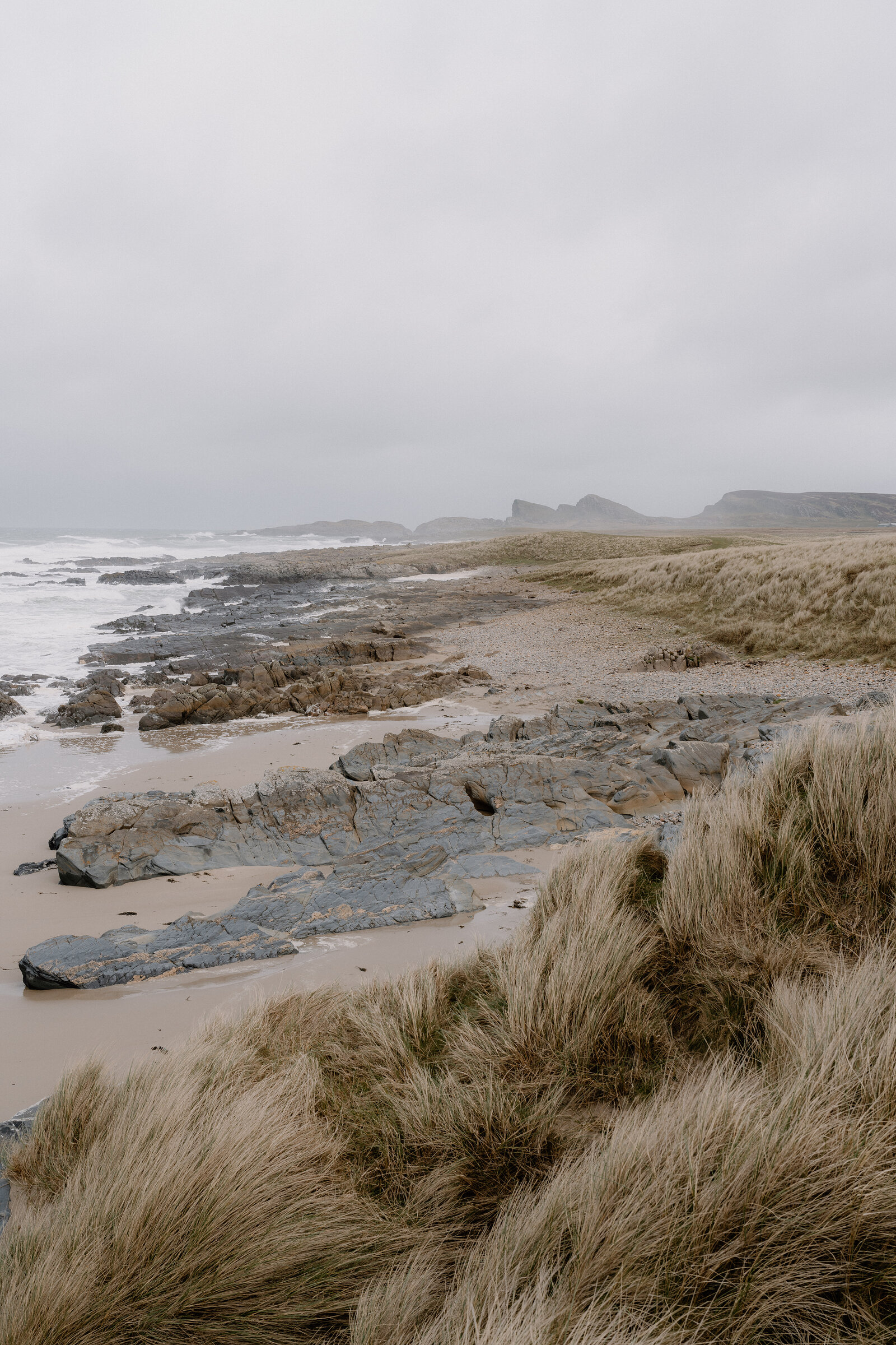 Grassy dunes slope down to a rocky shore where distant sea stacks rise from the water.