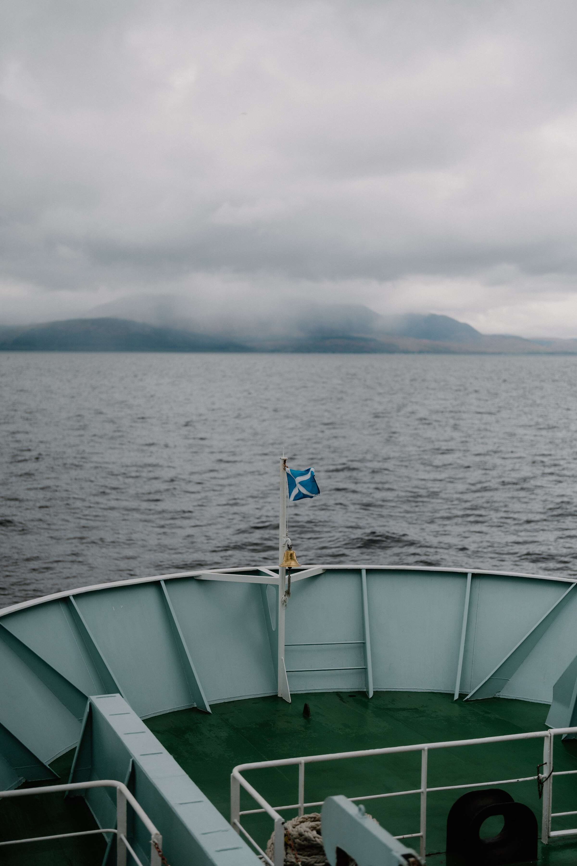 A ferry's blue and white Saltire flag flies from the bow as it crosses towards a mountainous island wreathed in low cloud.