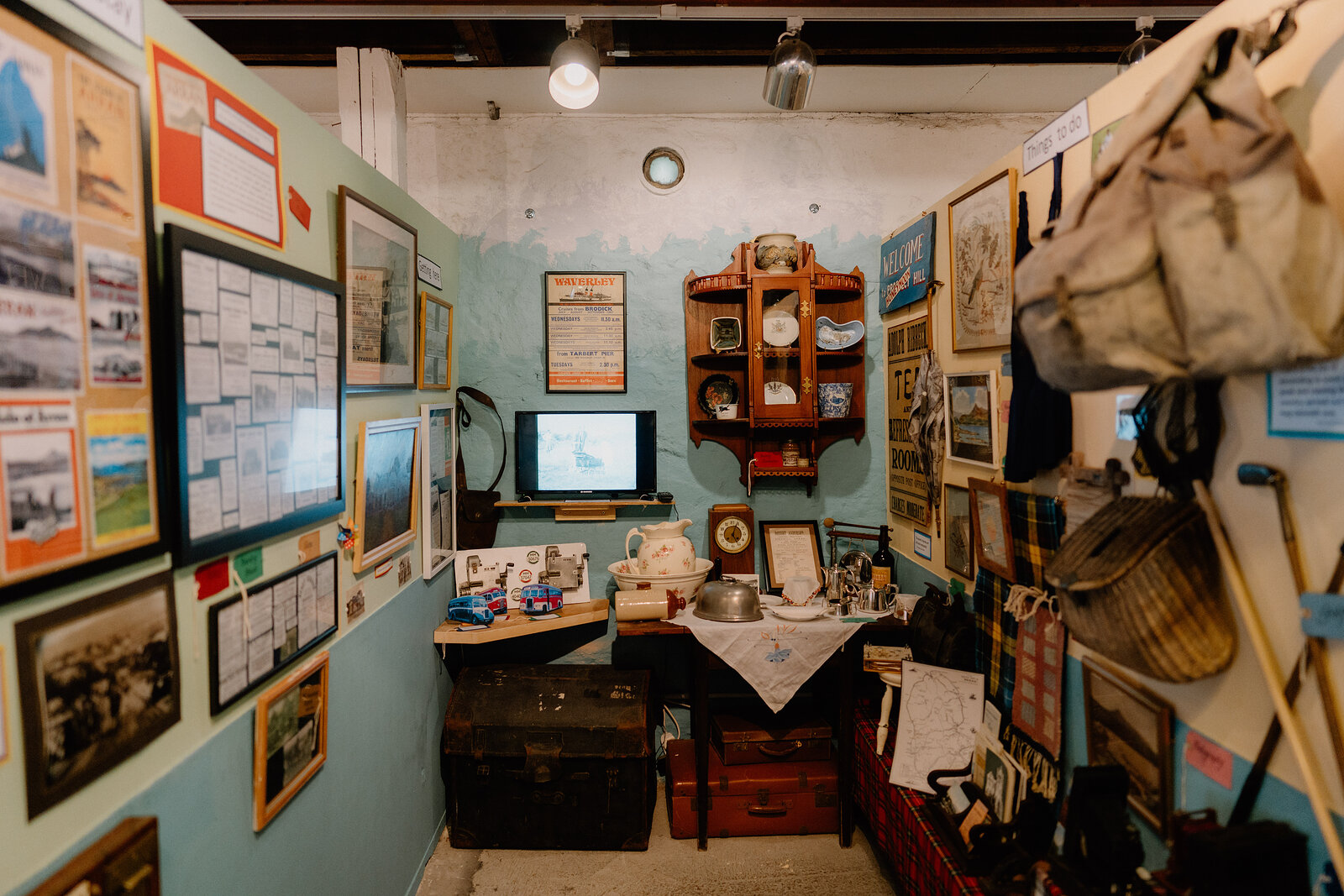 Framed photographs and documents cover the walls of a small museum room, a corner display cabinet and stacked trunks standing beside vintage sports equipment.