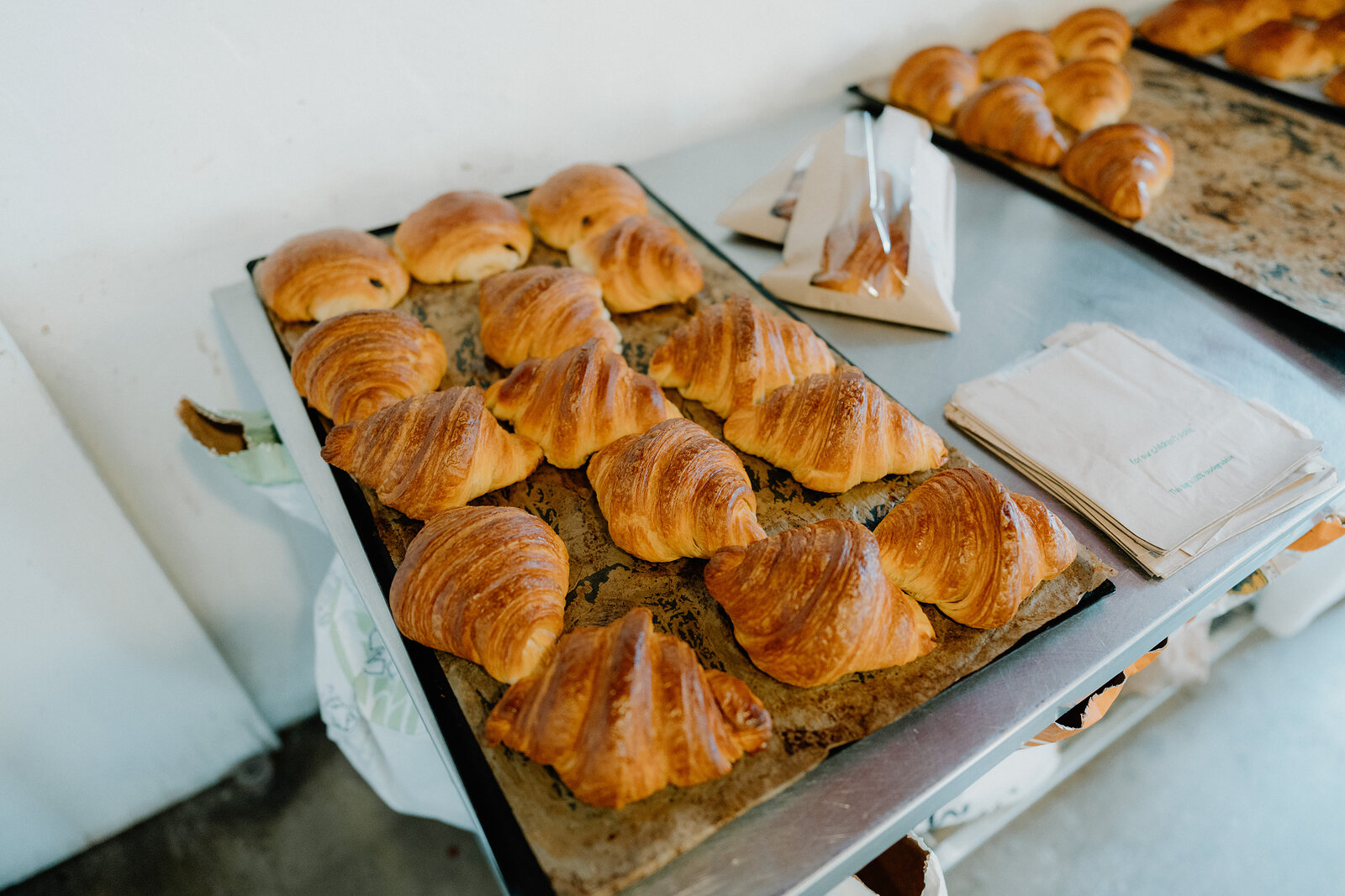 A tray of golden croissants sits on a baking sheet, a paper bag folded beside them.