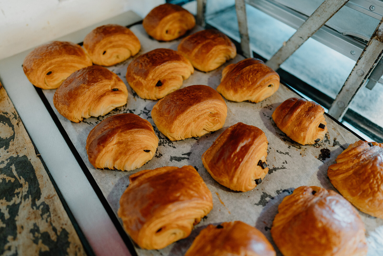 Rows of glazed pastries and chocolate-filled croissants sit on a baking tray in a bakery display cabinet.