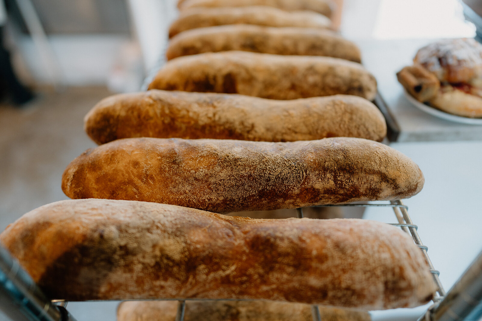 Long crusty loaves of bread cool in rows on a wire rack, a pastry visible on a plate beside them.