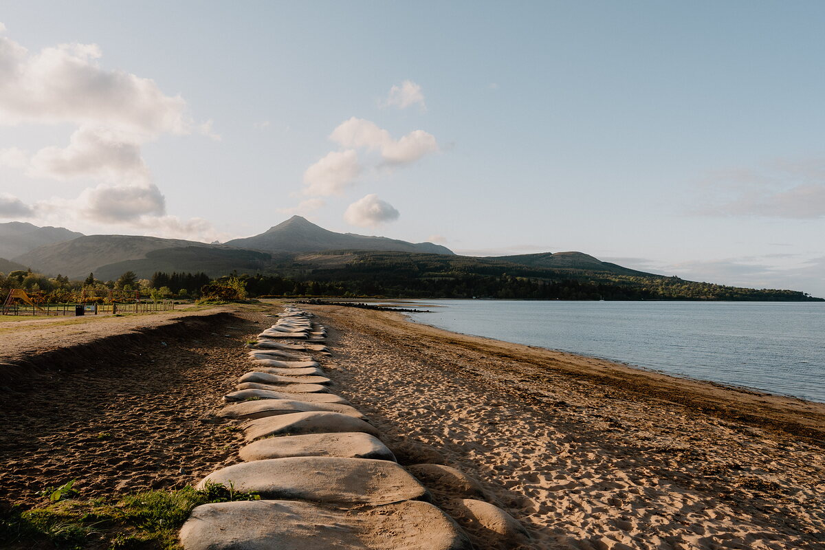 A line of stone breakwater blocks runs along a sandy beach at sunrise, a conical mountain peak rising beyond the trees.
