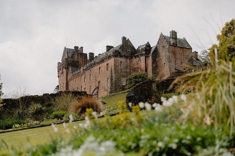 A long red sandstone castle with crow-stepped gables and chimneys stands above terraced garden borders in bloom.