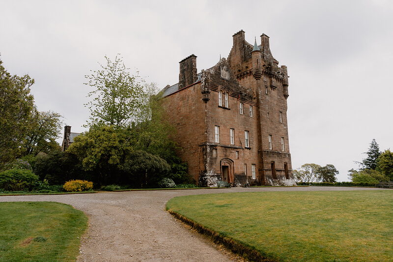 A tall red sandstone tower house with crow-stepped gables and dormer windows stands beyond a curving gravel path and lawn.