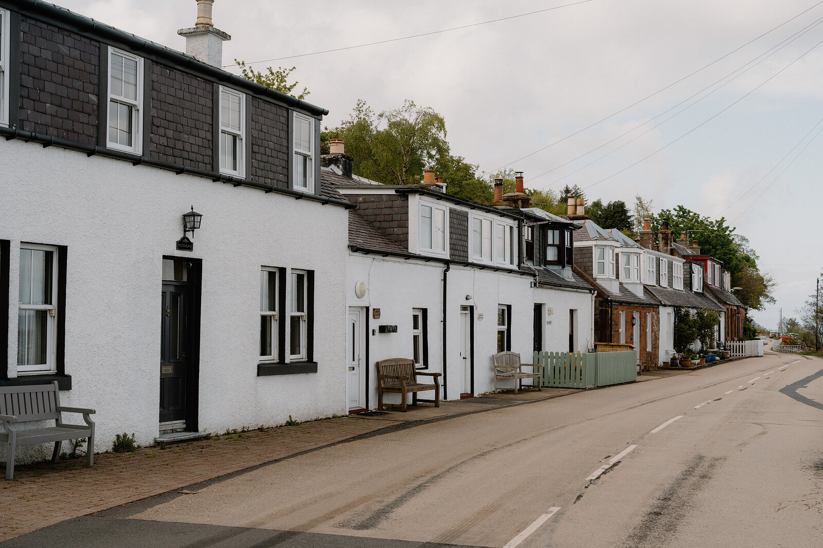 A row of white and stone cottages with dormer windows lines a quiet coastal street, benches set outside two of the doors.
