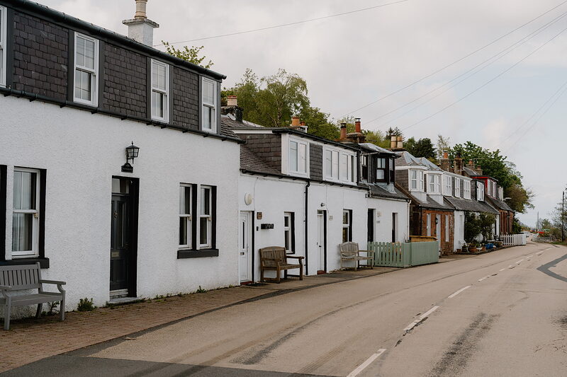 A row of white and stone cottages with dormer windows lines a quiet coastal street, benches set outside two of the doors.
