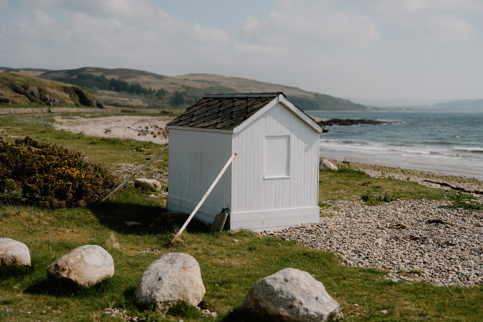 A white timber beach hut with a slate roof stands propped with a wooden strut among boulders on a grassy shore.