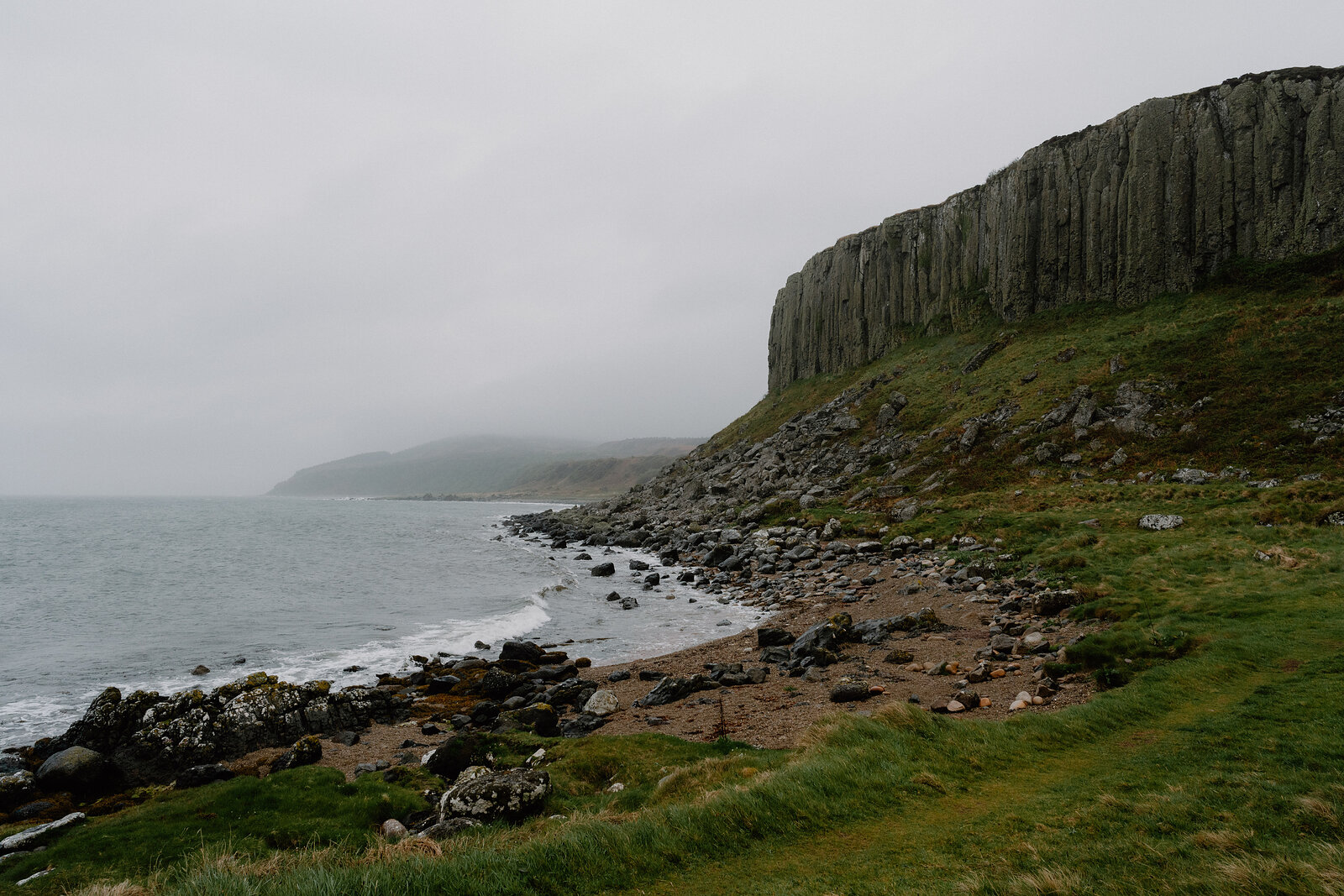 Columned basalt cliffs rise above a pebble and rock shore where waves break gently, low hills fading into mist along the coastline.