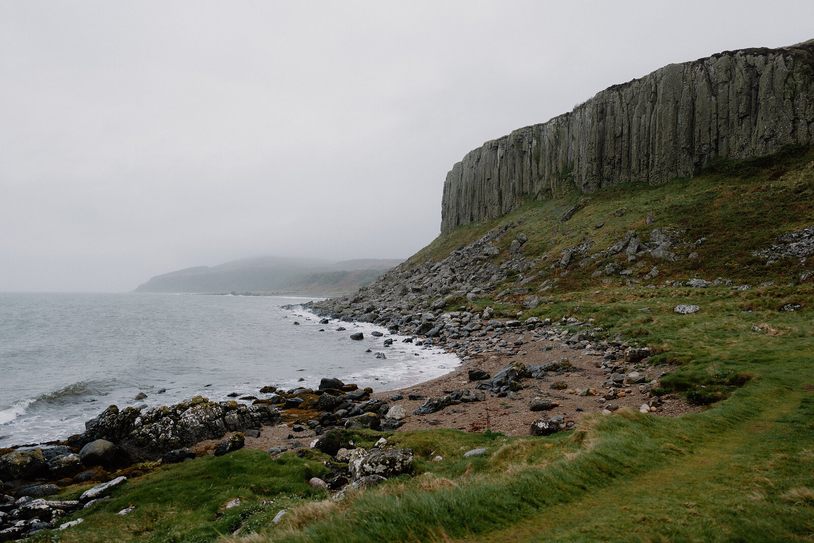 Columned basalt cliffs rise above a pebble shore beside a calm sea, low hills fading into mist along the coastline.