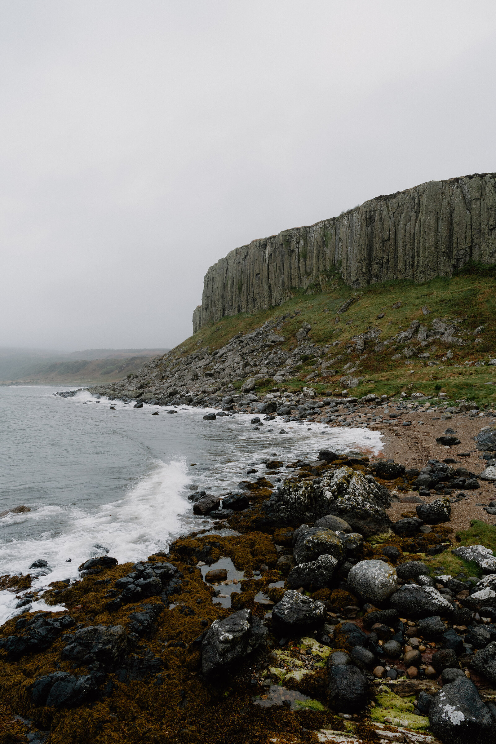 Waves break over seaweed-covered rocks at the base of columned basalt cliffs beneath an overcast sky.