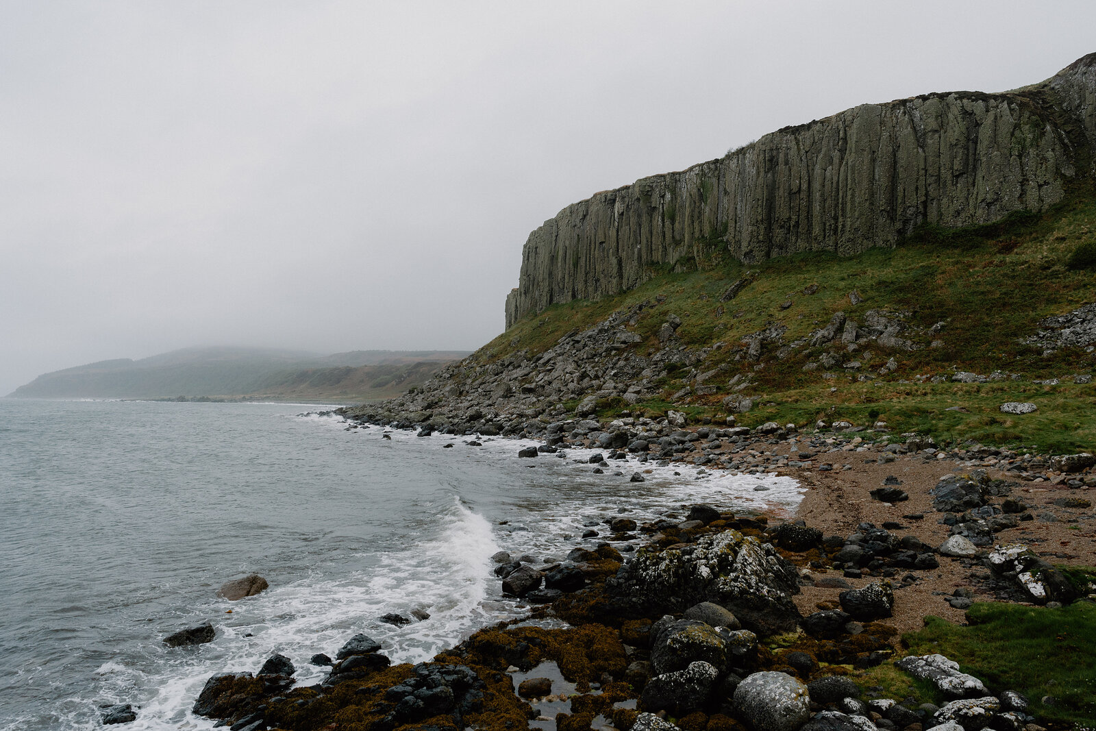 Columned basalt cliffs rise above a rocky, seaweed-strewn shore where gentle waves break beneath a misty sky.