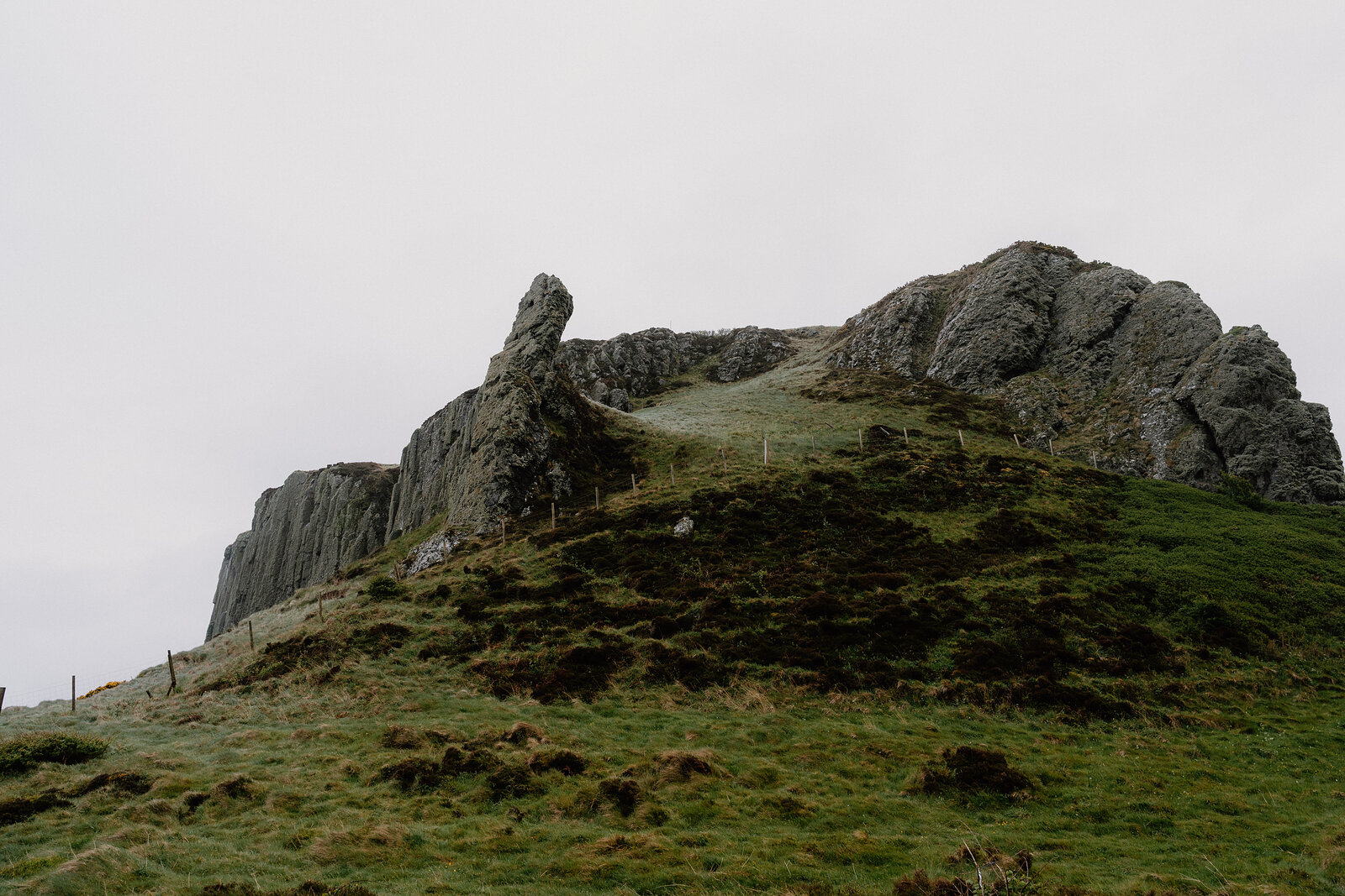 A jagged rock pinnacle rises from a grassy, moss-covered ridge beside a rocky outcrop under an overcast sky.