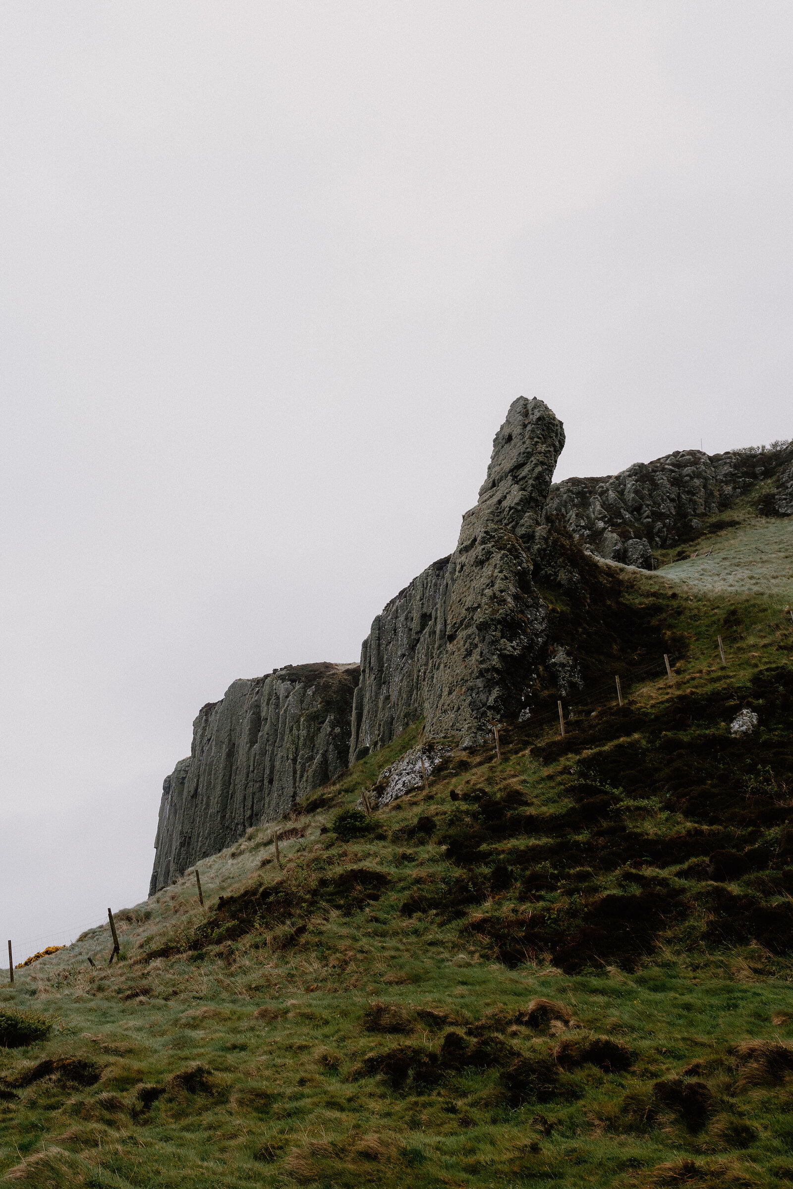 A jagged rock pinnacle rises from a grassy, fenced hillside beside columned cliffs under an overcast sky.