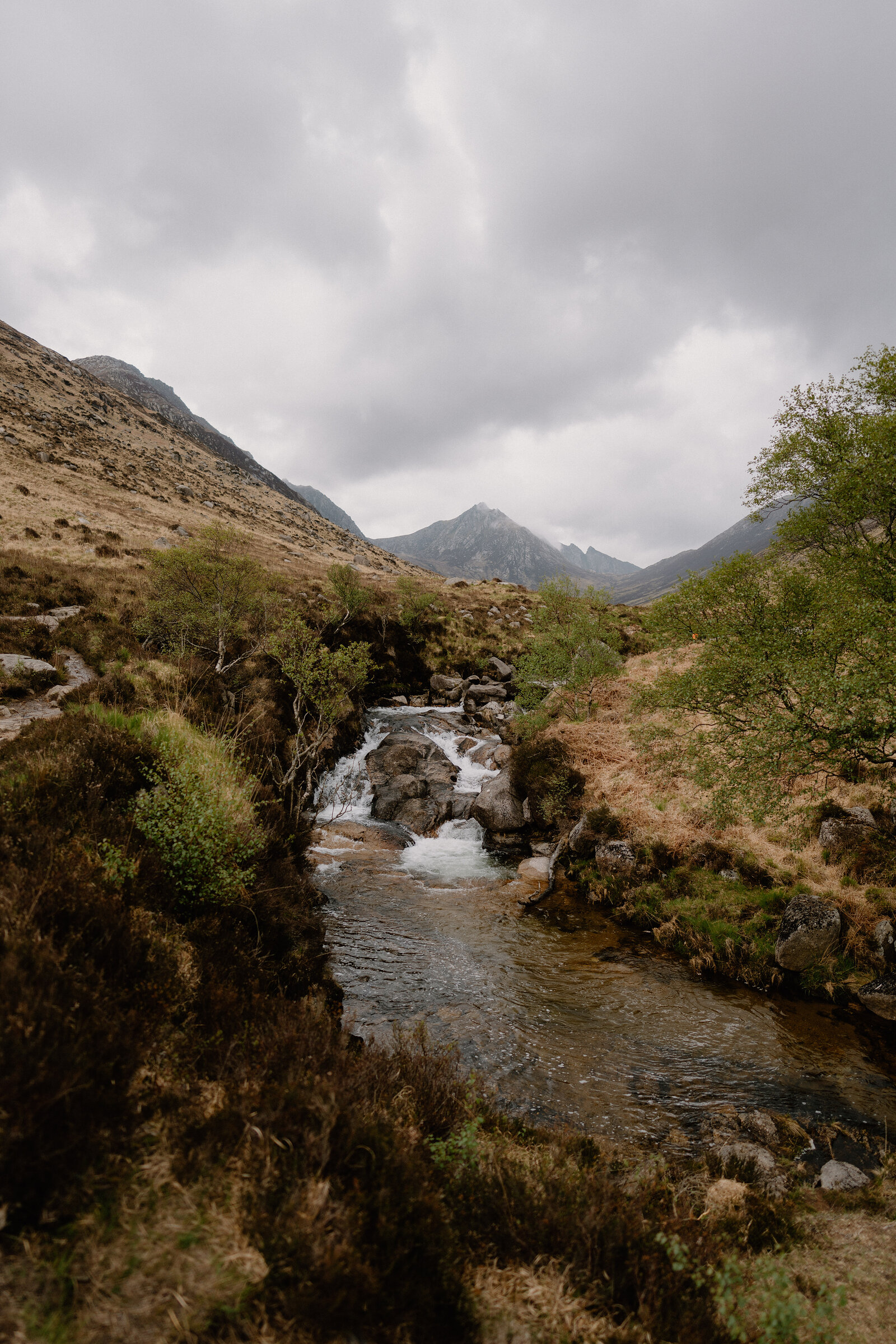 A river tumbles over rock ledges into a pool at the foot of a mountain glen, jagged peaks rising ahead under a cloudy sky.