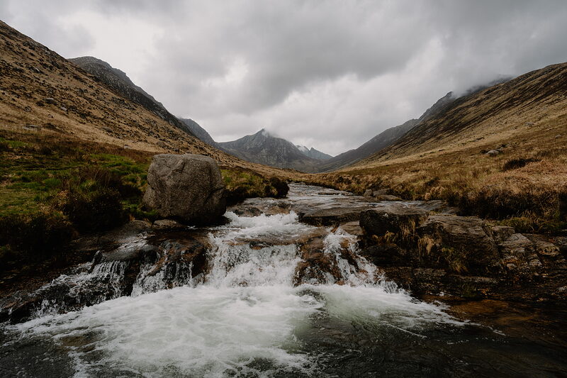 A river rushes over rocks past a large boulder in a mountain glen, snow-dusted peaks rising at the head of the valley.