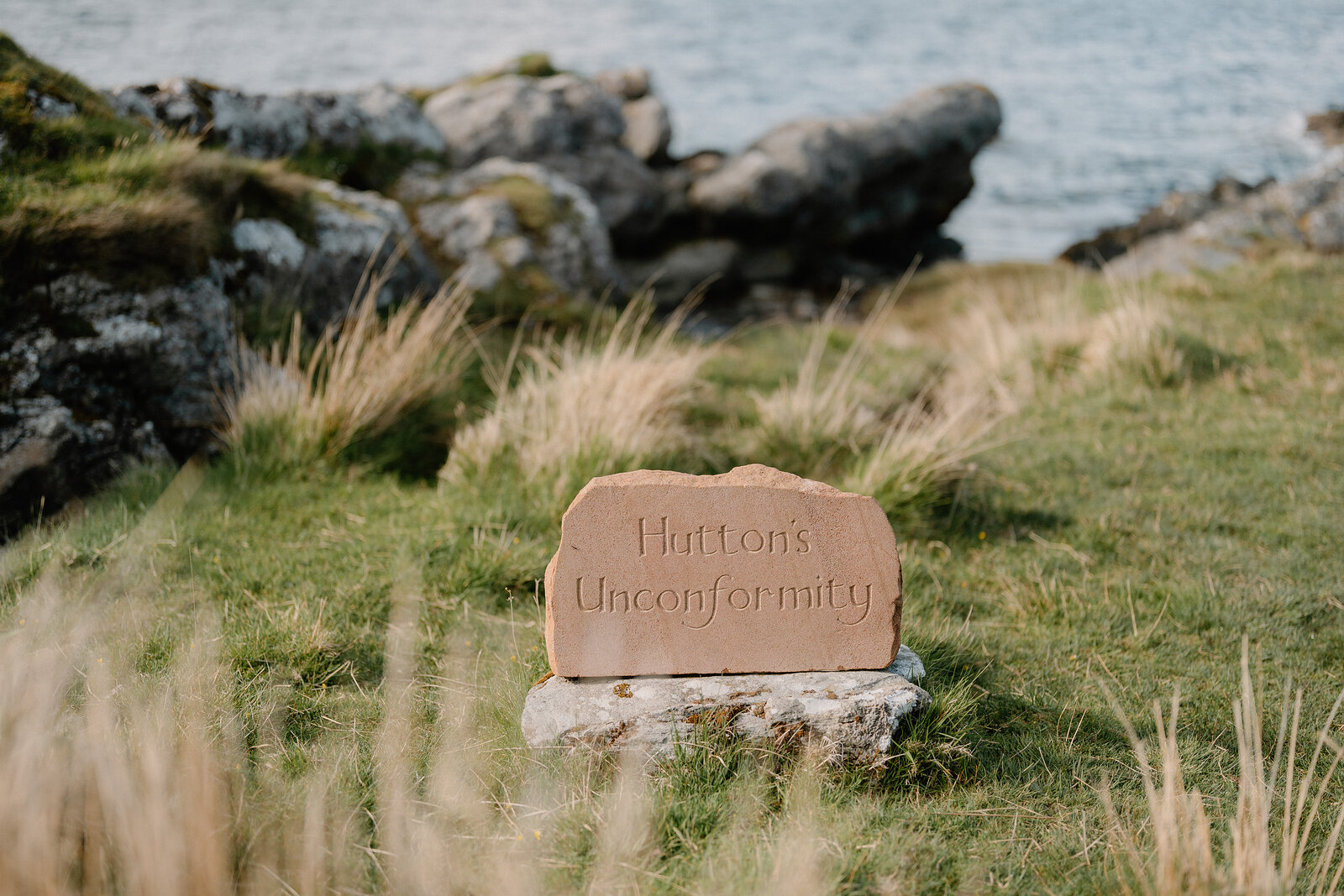 A carved sandstone plaque reading 'Hutton's Unconformity' sits on a boulder among long grass above a rocky shoreline.