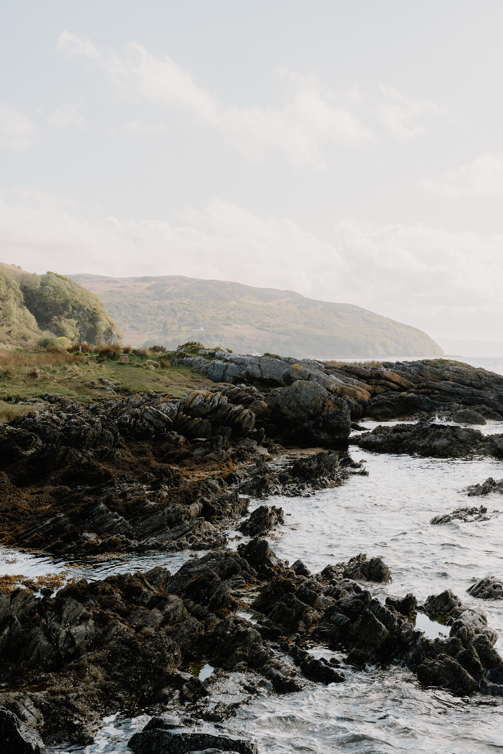 Lichen-streaked rocks line a grassy shoreline, a wooded headland curving into the sea beyond.
