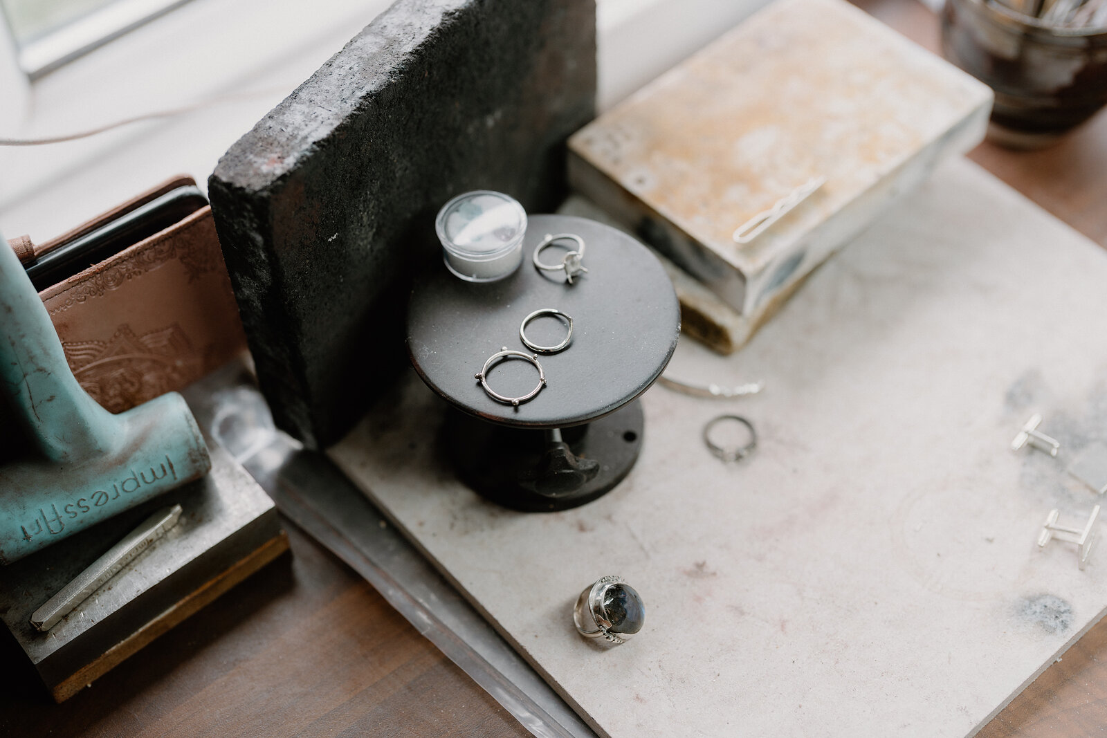 Silver rings sit on a black stand beside a hammer and books on a jeweller's workbench.