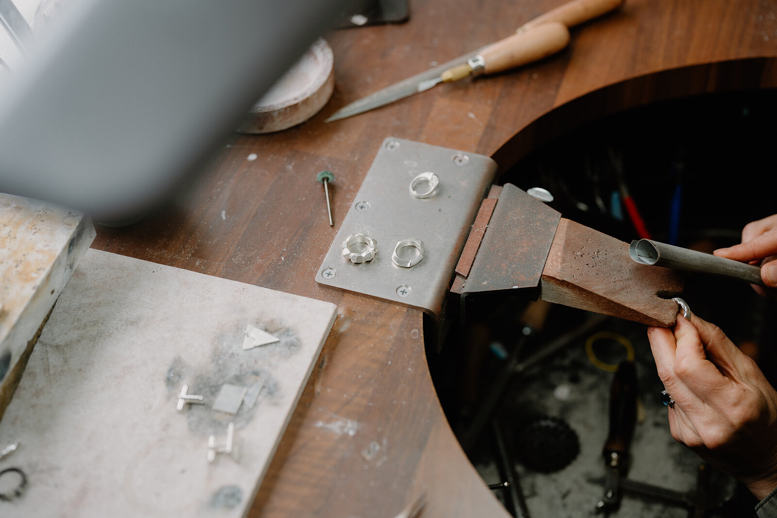 A hand holds pliers to shape a small metal ring clamped in a vice, silver rings and tools scattered across the workbench.