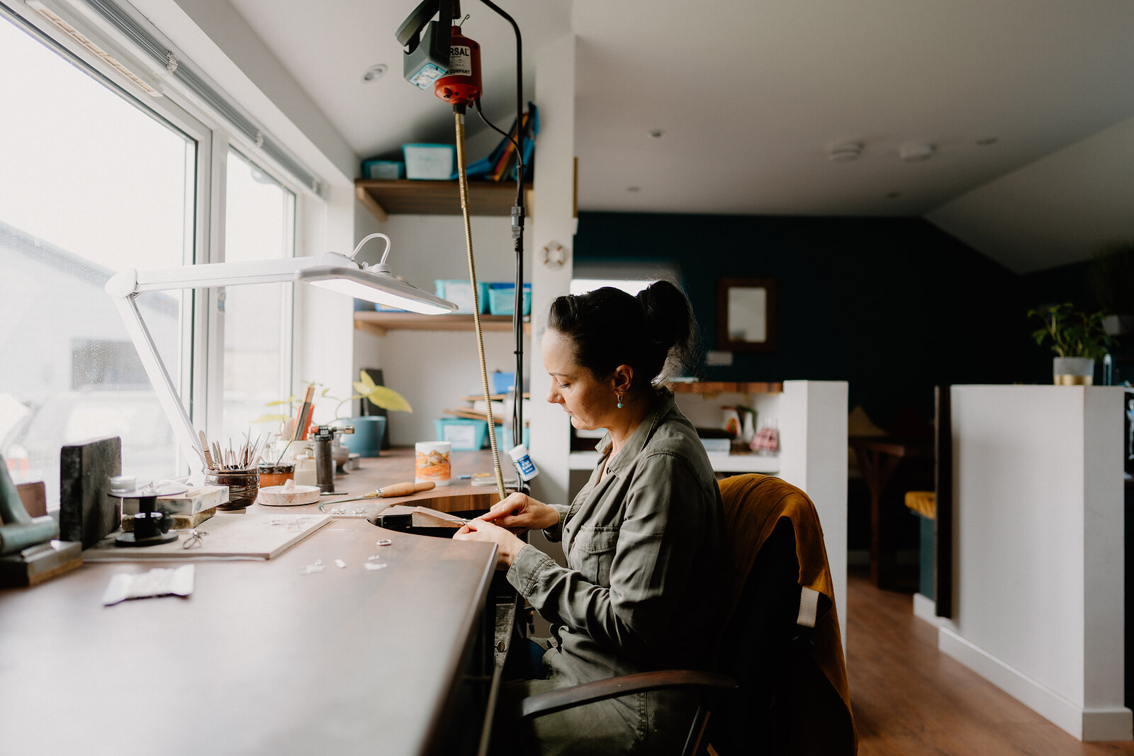 A woman sits at a jeweller's workbench beneath a desk lamp, tools and small stones arranged in front of her, a kitchen visible behind.