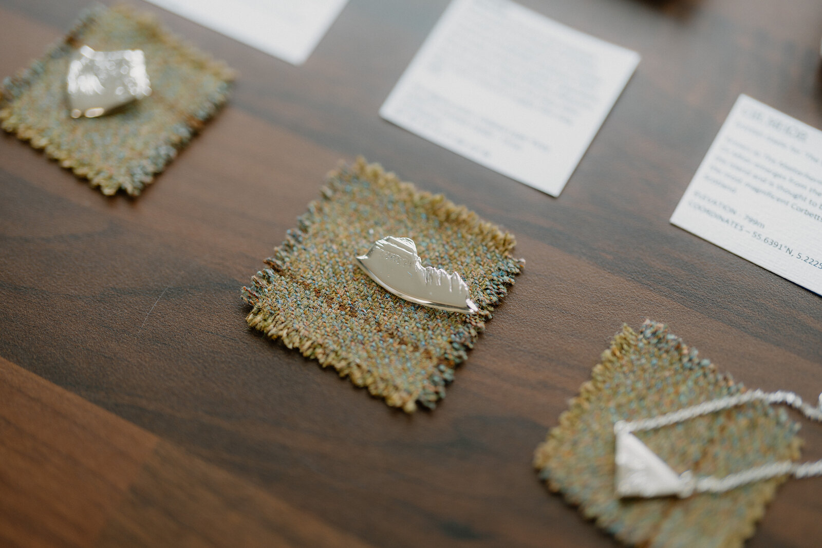 Small bird and leaf-shaped silver brooches sit on hessian squares beside information cards on a wooden table.